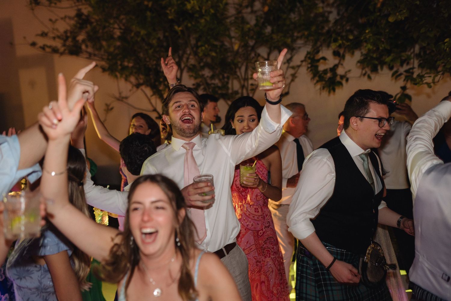 Party guests dancing enthusiastically with raised arms on a crowded dance floor with warm lighting.