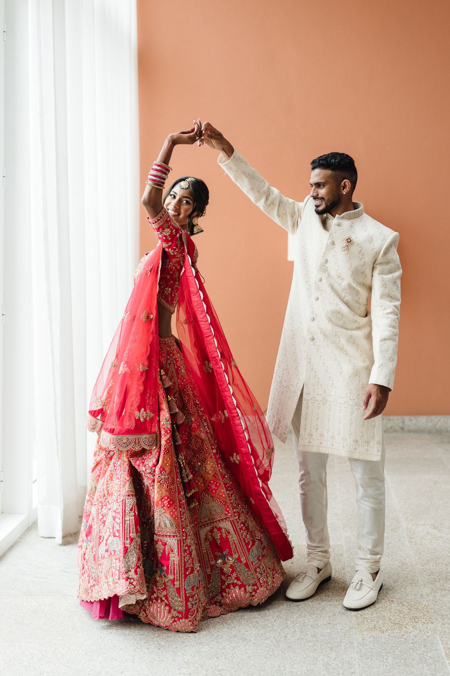 Couple in traditional Indian wedding attire posing playfully against a peach-colored wall.