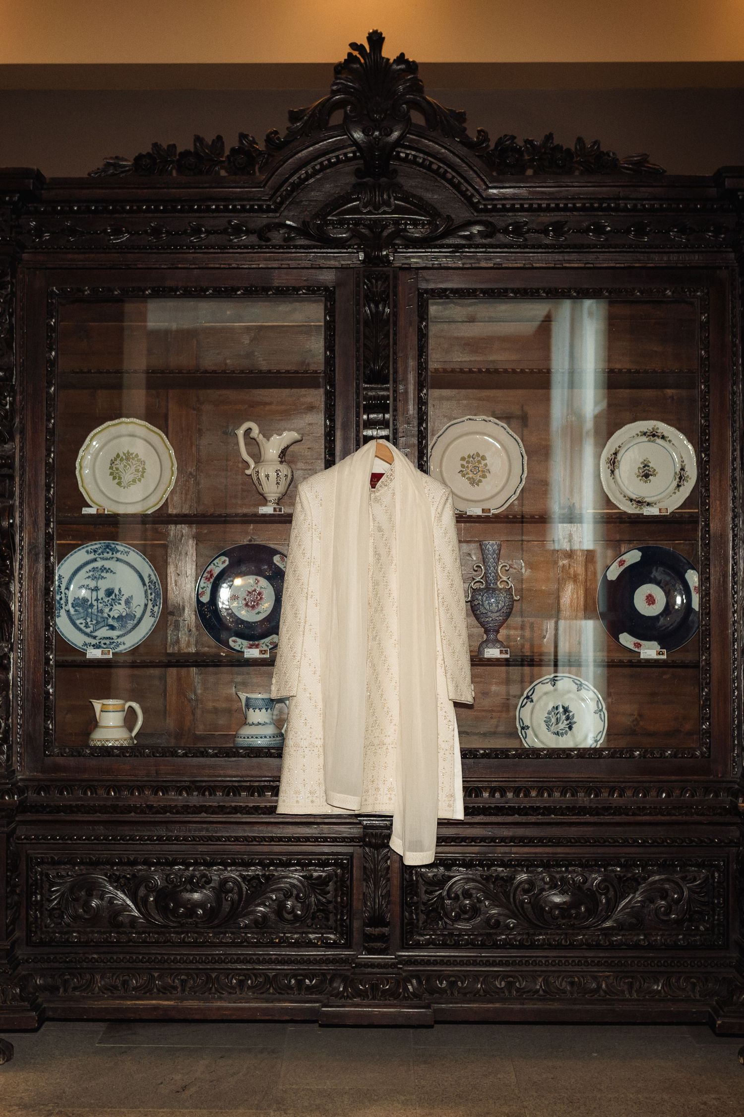 Ornate wooden display cabinet with antique plates and white garment hanging in front.