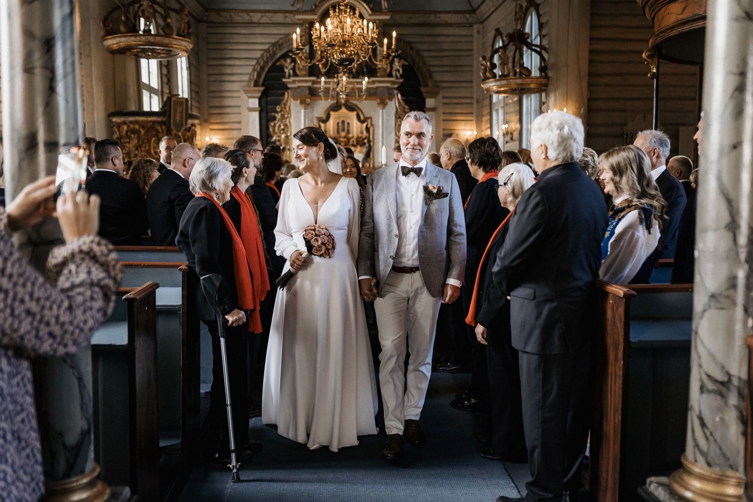 Wedding guests line the aisle of a historic church with elegant chandeliers overhead.
