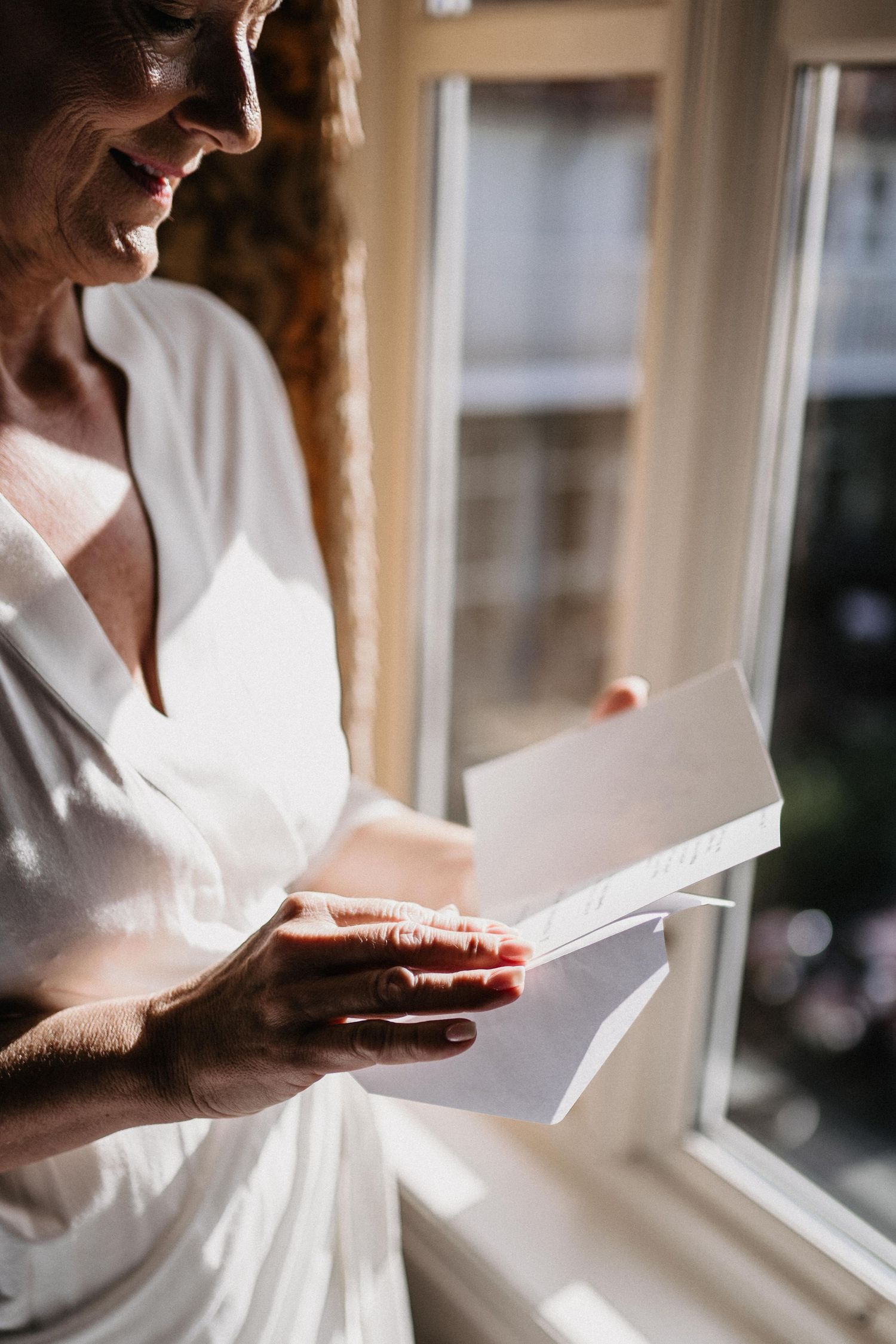 Someone in a white dress reads papers while standing by a window.