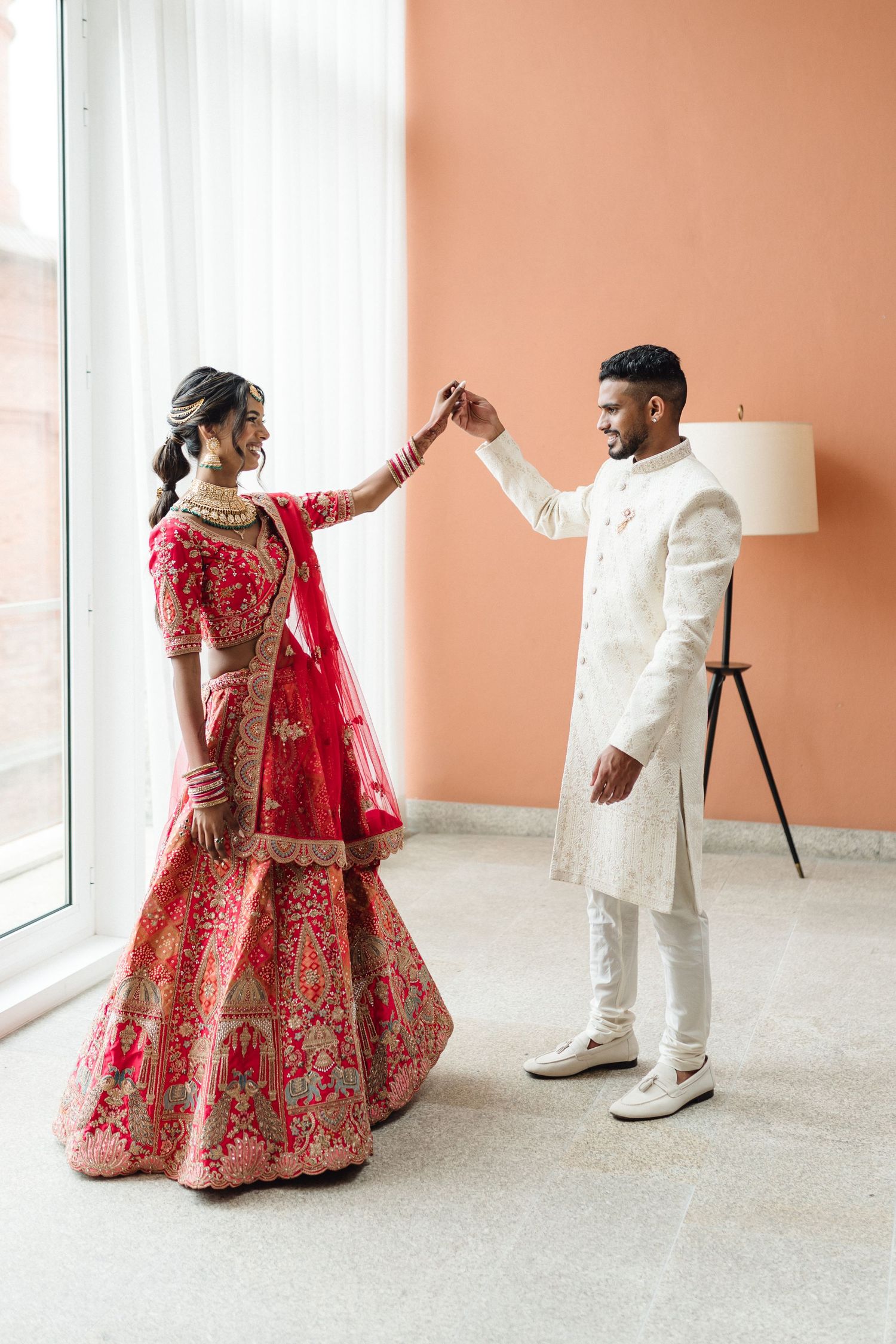 Wedding couple in red lehenga and white sherwani dance in elegant indoor setting.