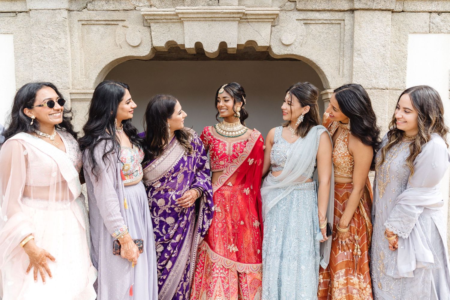 Wedding party members in colorful traditional attire pose against a stone architectural backdrop.