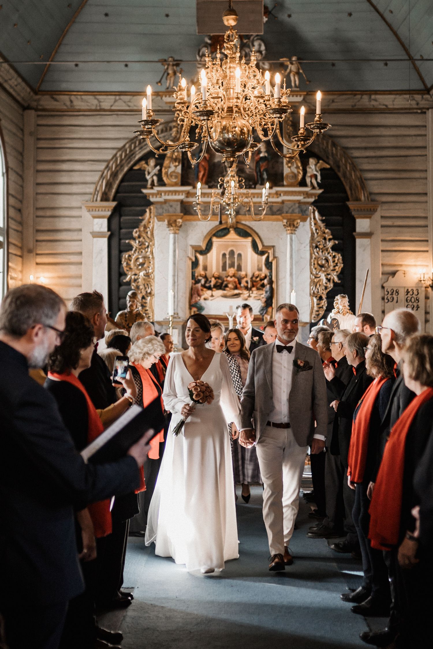Wedding guests gather in an elegant ballroom under ornate crystal chandeliers during a celebration.