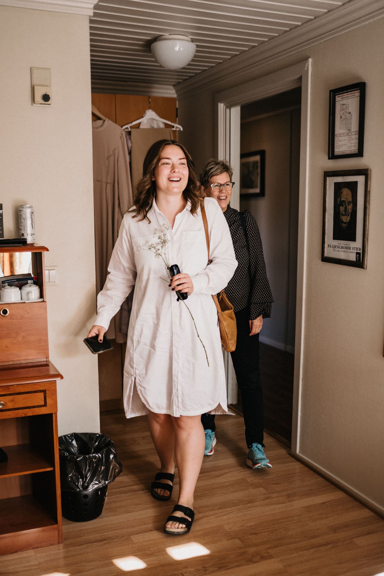 Person walking down a hallway wearing a white shirt and dark sandals.