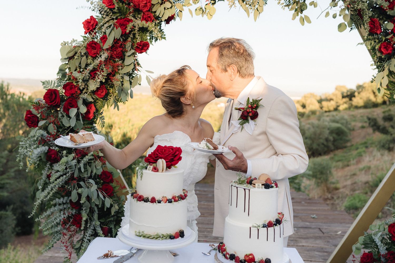 Wedding celebration under floral arch with red roses and greenery at sunset.