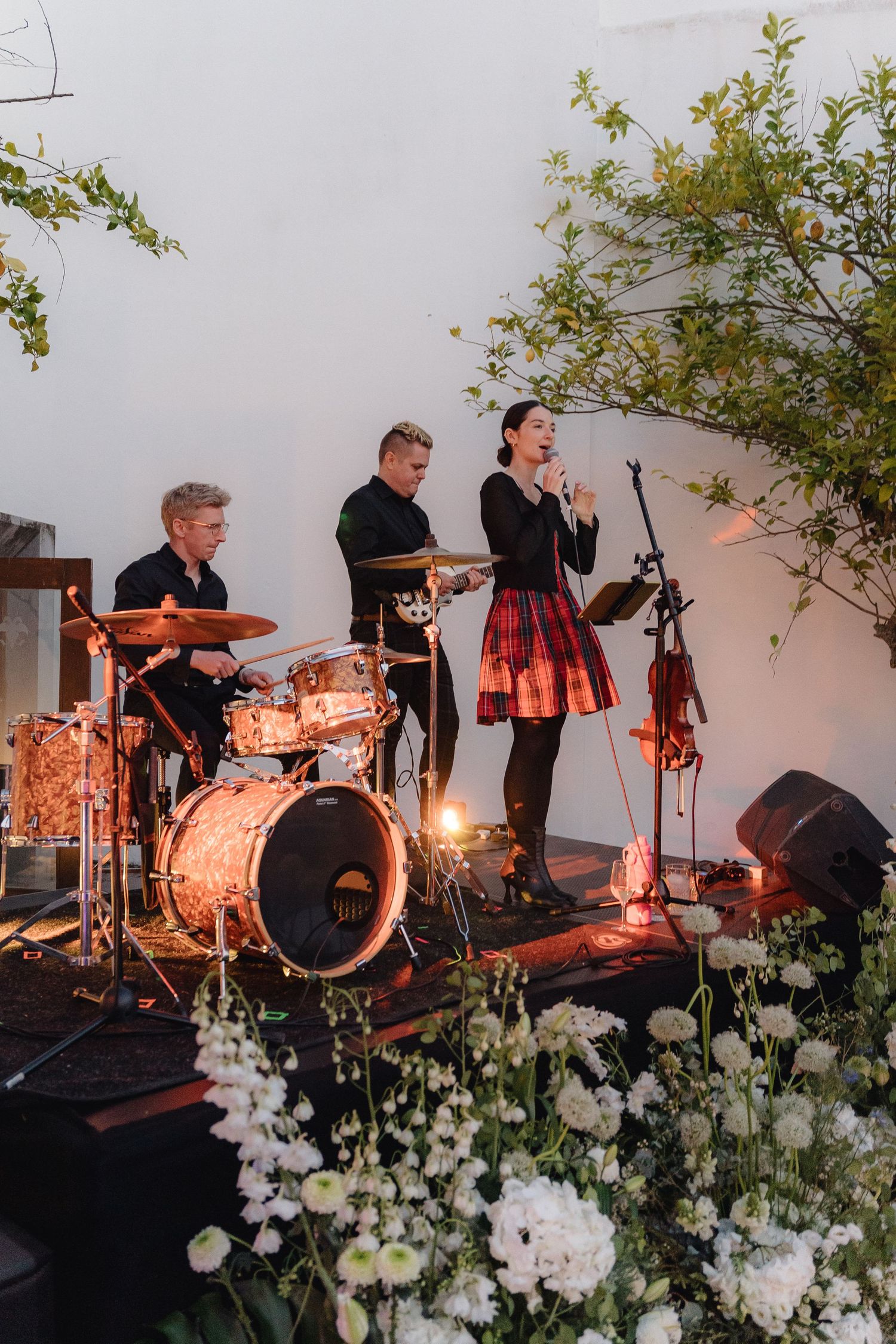 Band performs on stage with drums and instruments surrounded by white floral arrangements.