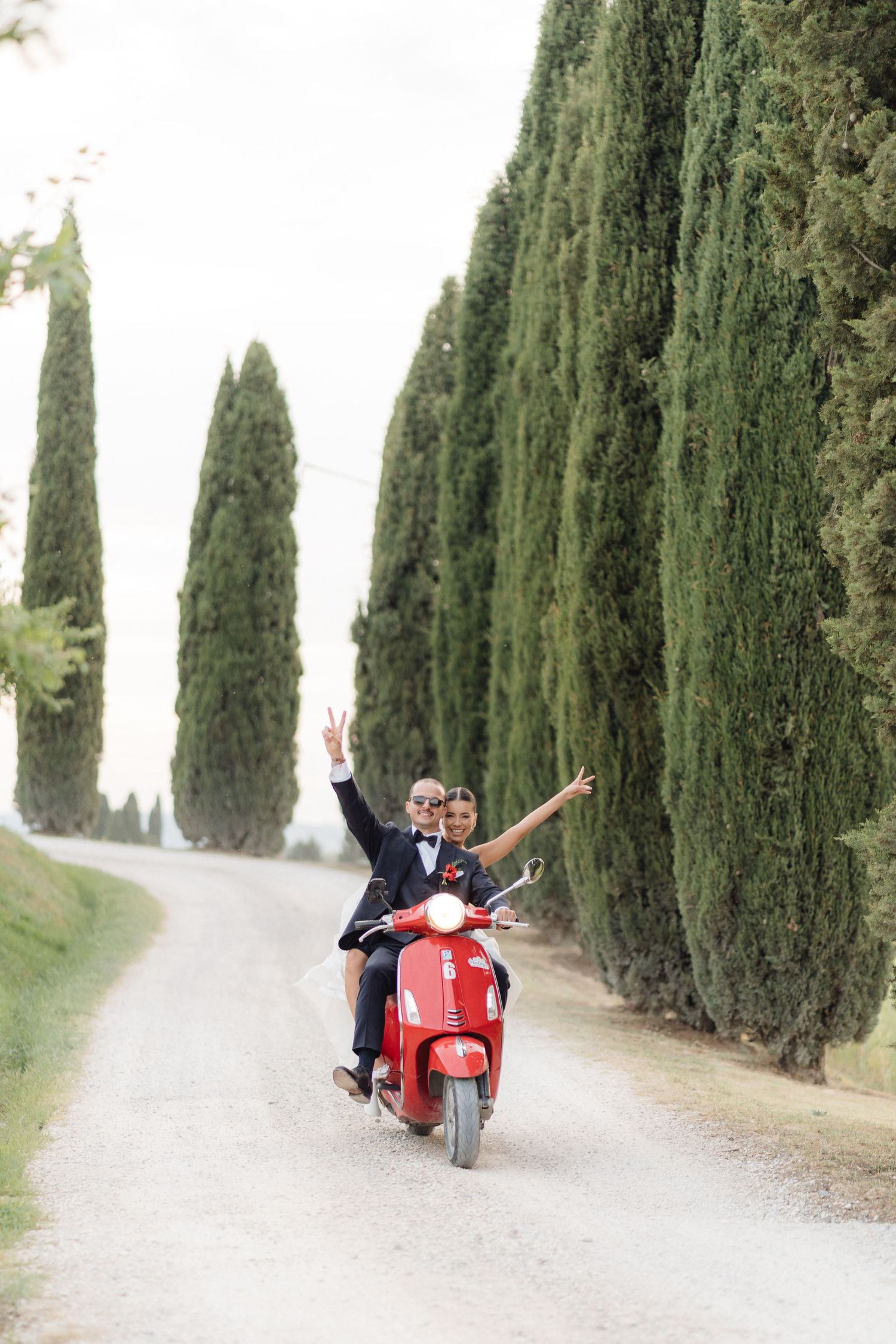 Joyful newlyweds riding red Vespa scooter down cypress tree-lined gravel road in Tuscany, Italy.