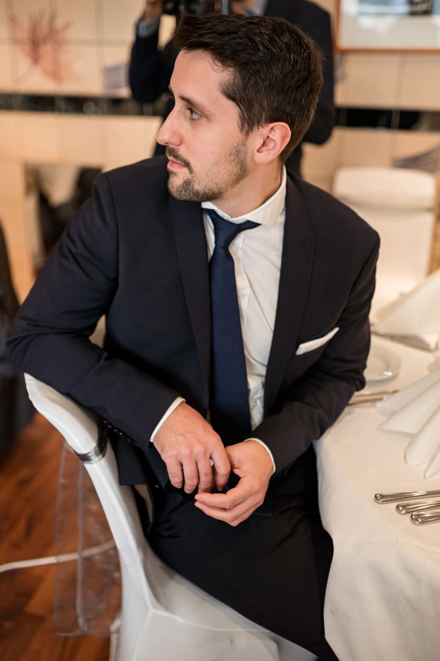Groom in navy suit relaxing on white furniture during wedding reception.