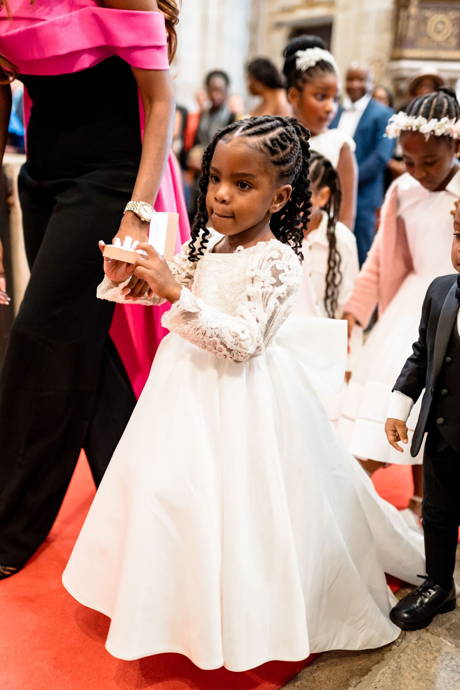 Young attendants in white dresses participate in a formal wedding ceremony on a red carpet.