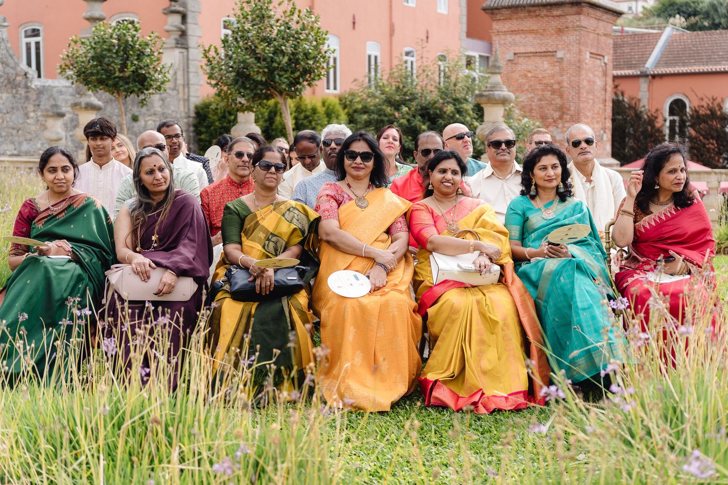 Large group photo of wedding guests in colorful traditional Indian attire sitting outdoors on grass.