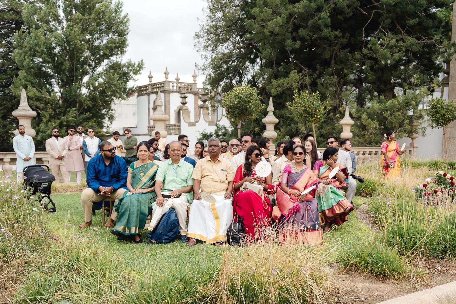 Wedding guests seated outdoors on chairs in a garden setting with ornate architecture in the background.