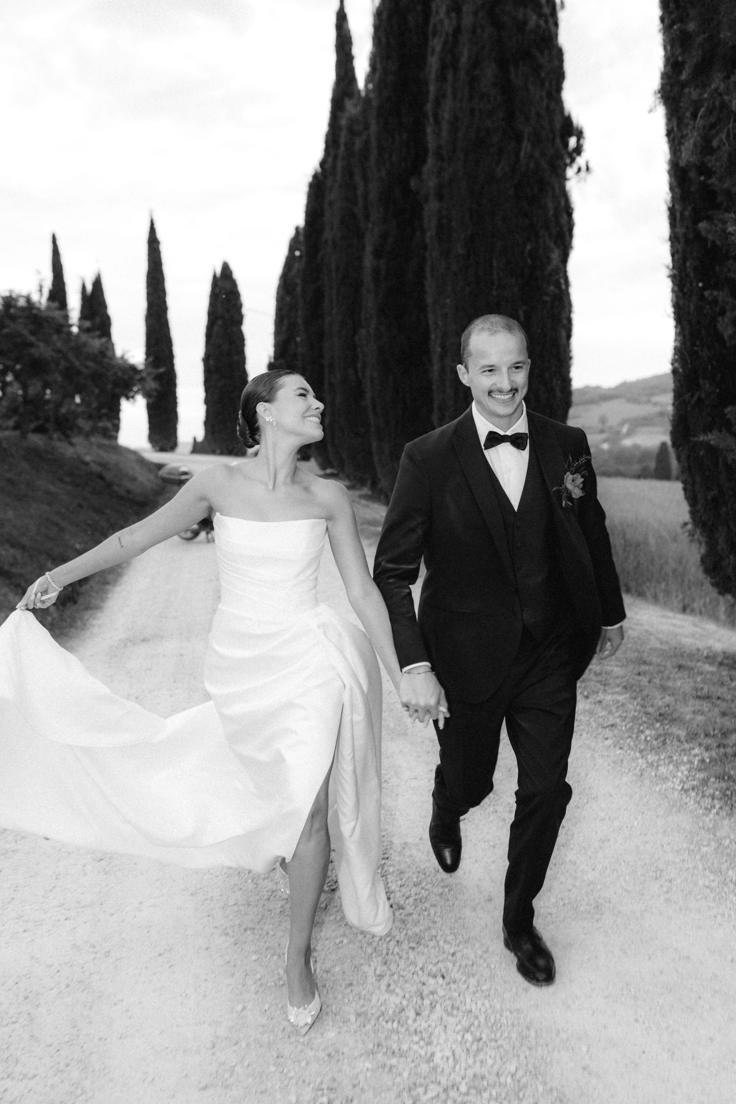 Bride and groom holding hands walking down cypress tree lined path in black and white wedding photo.