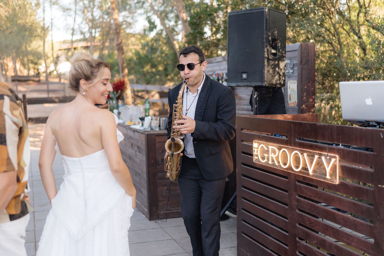 Saxophone player performs at outdoor wedding reception near wooden DJ booth.