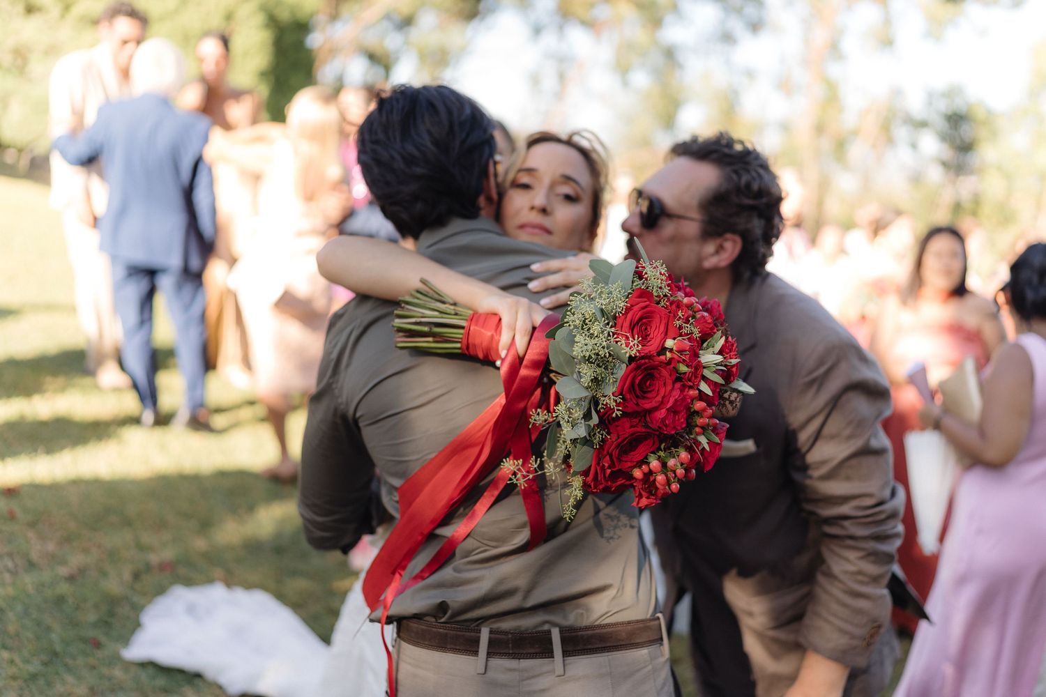 Wedding guests embrace while holding vibrant red bouquets at an outdoor ceremony.