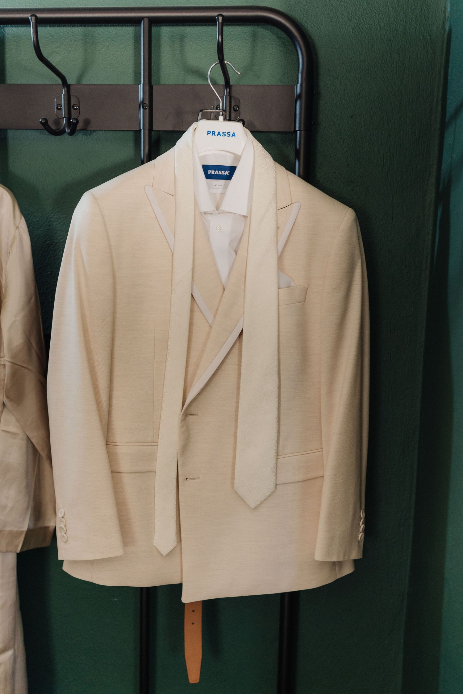 Cream colored wedding suits with ties hanging neatly on metal rack against green wall.
