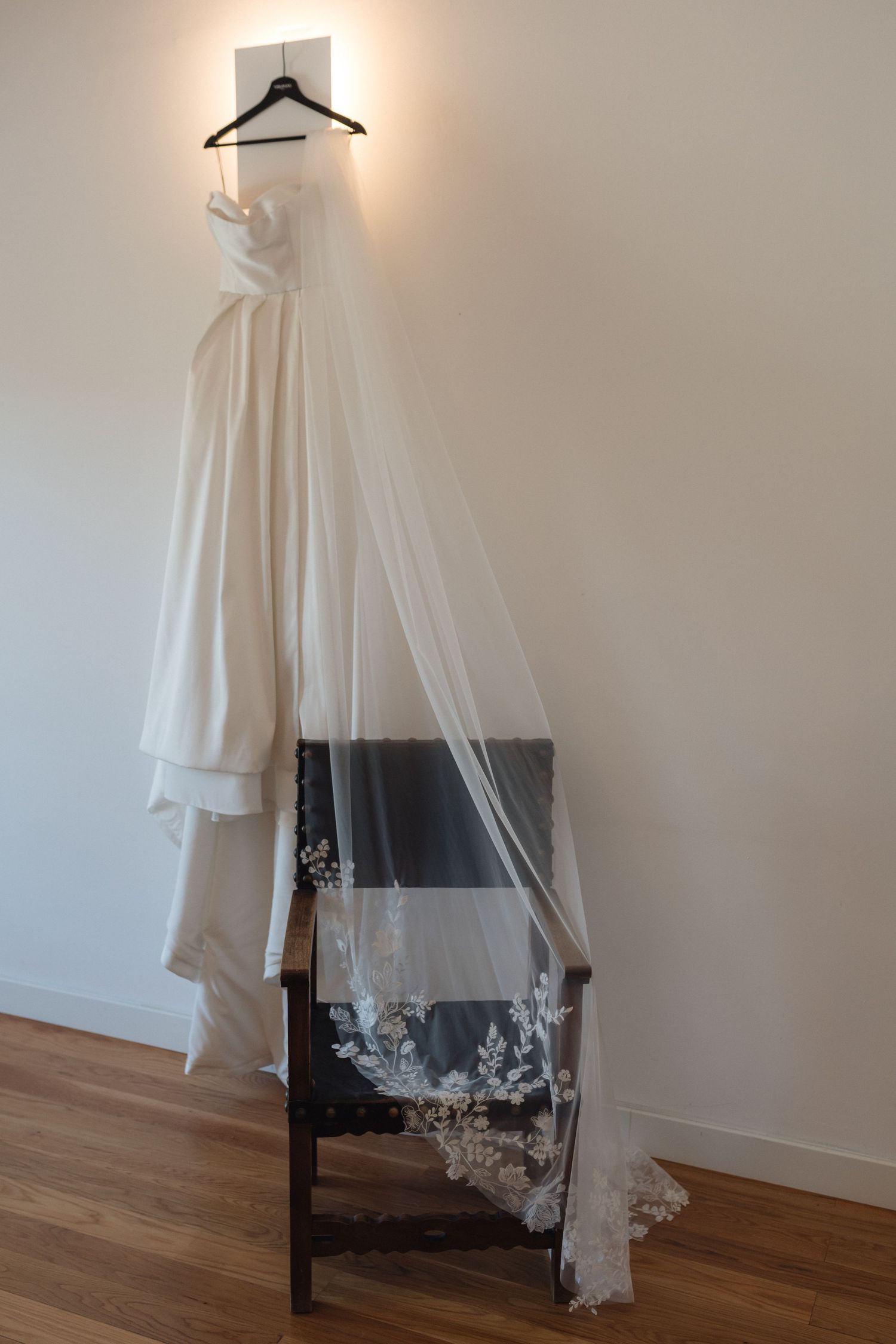 Elegant white wedding dress with lace veil hanging on display against neutral wall.
