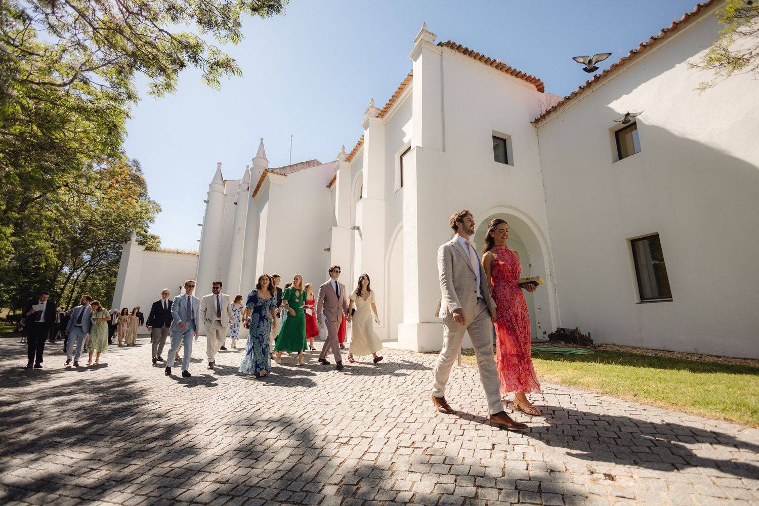 People walk along a white-walled pathway outside traditional Spanish colonial style buildings in bright sunlight.
