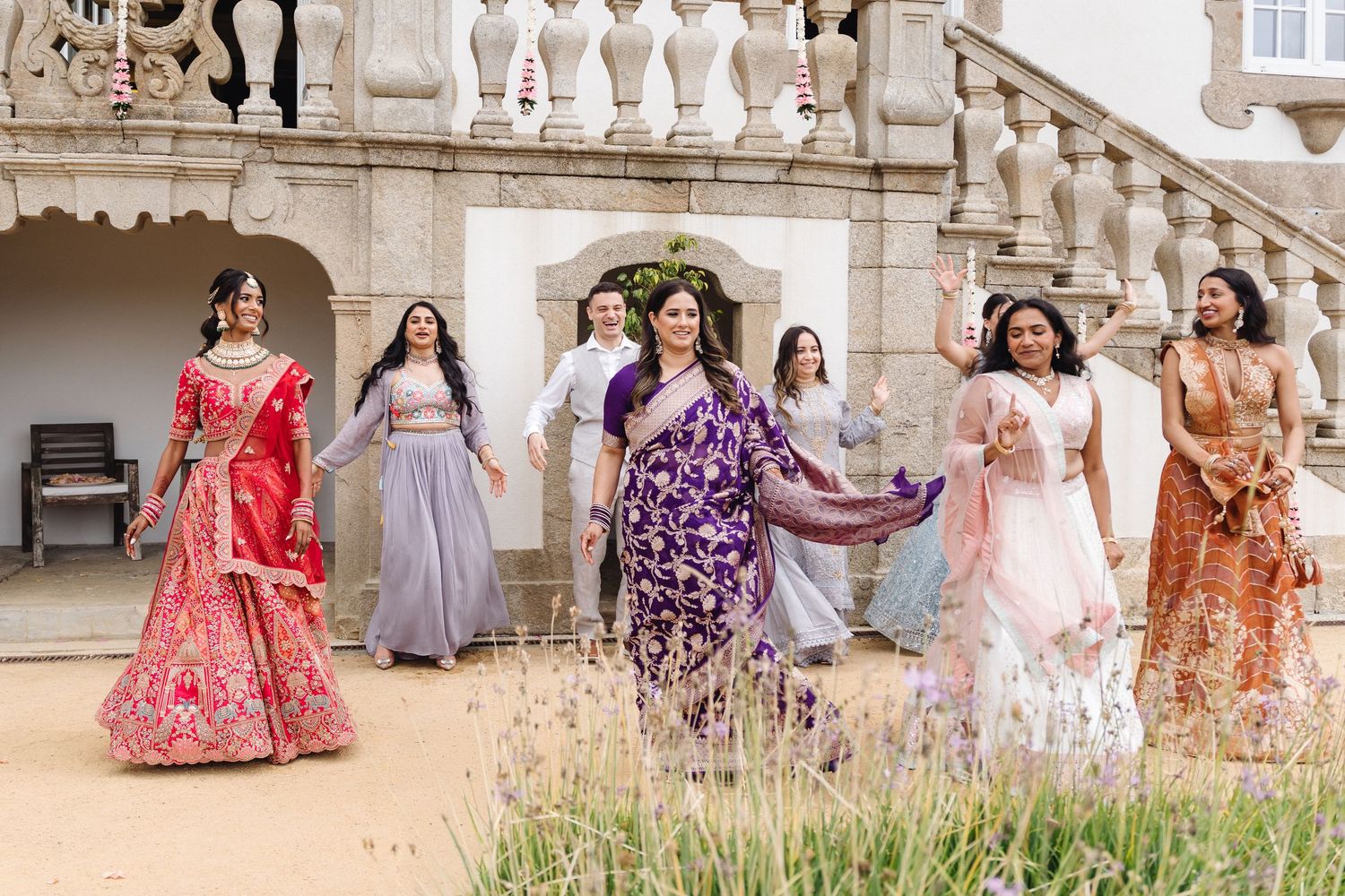 Wedding party lineup in colorful traditional and western attire against white stone building facade.