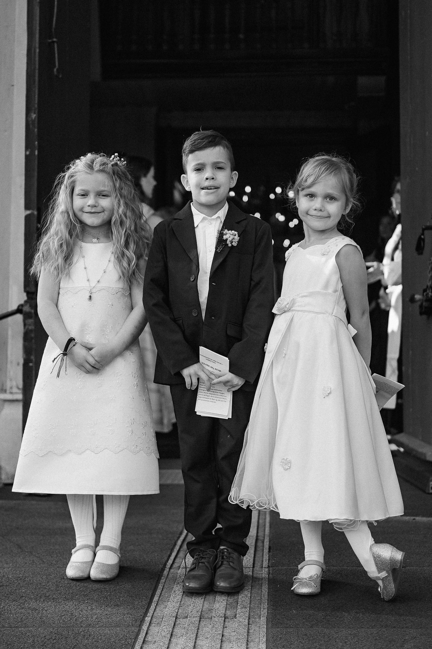 Row of children in formal white dresses and suits standing against dark background.
