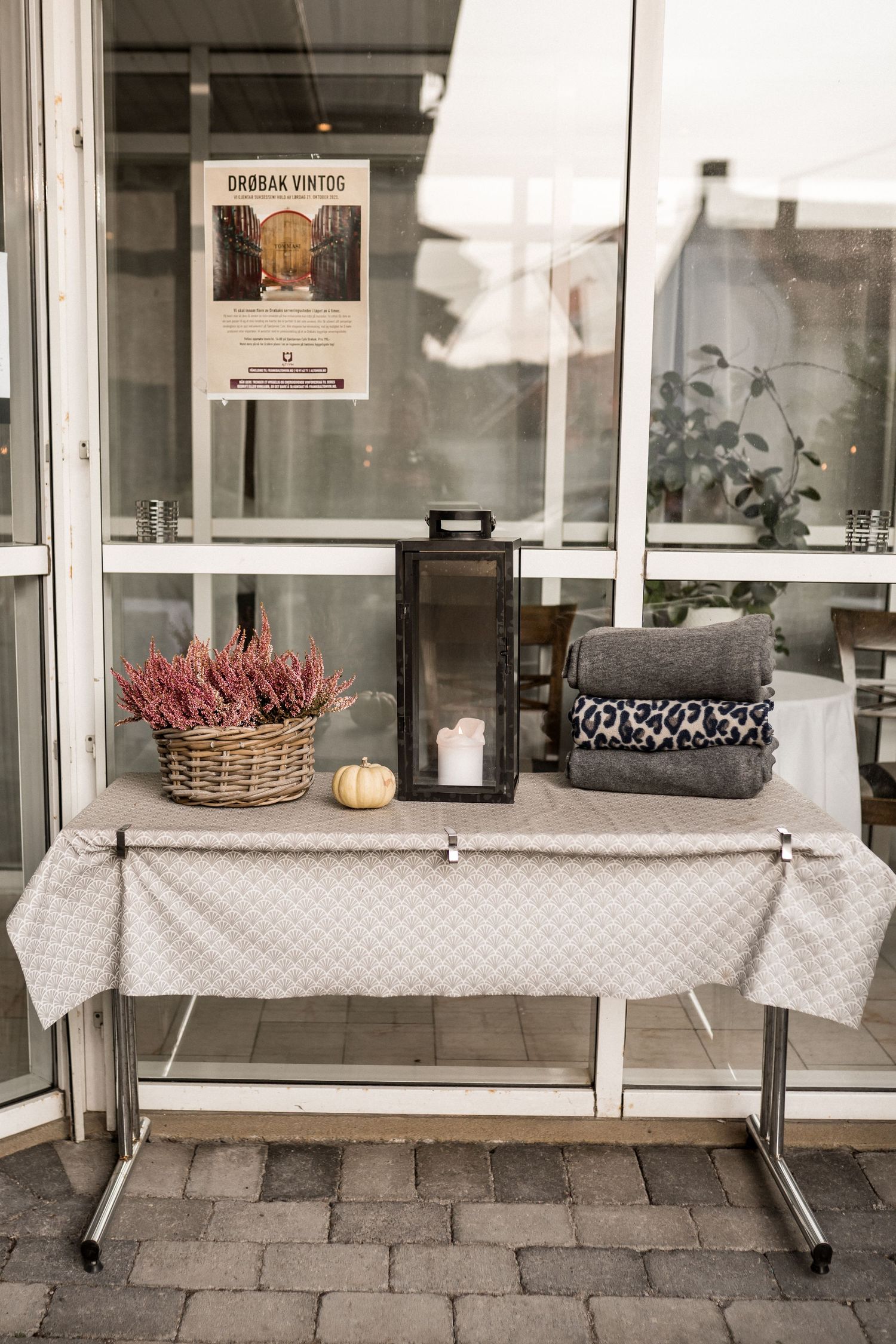 White tablecloths drape over benches with decorative heather plants and lanterns outside a glass building.