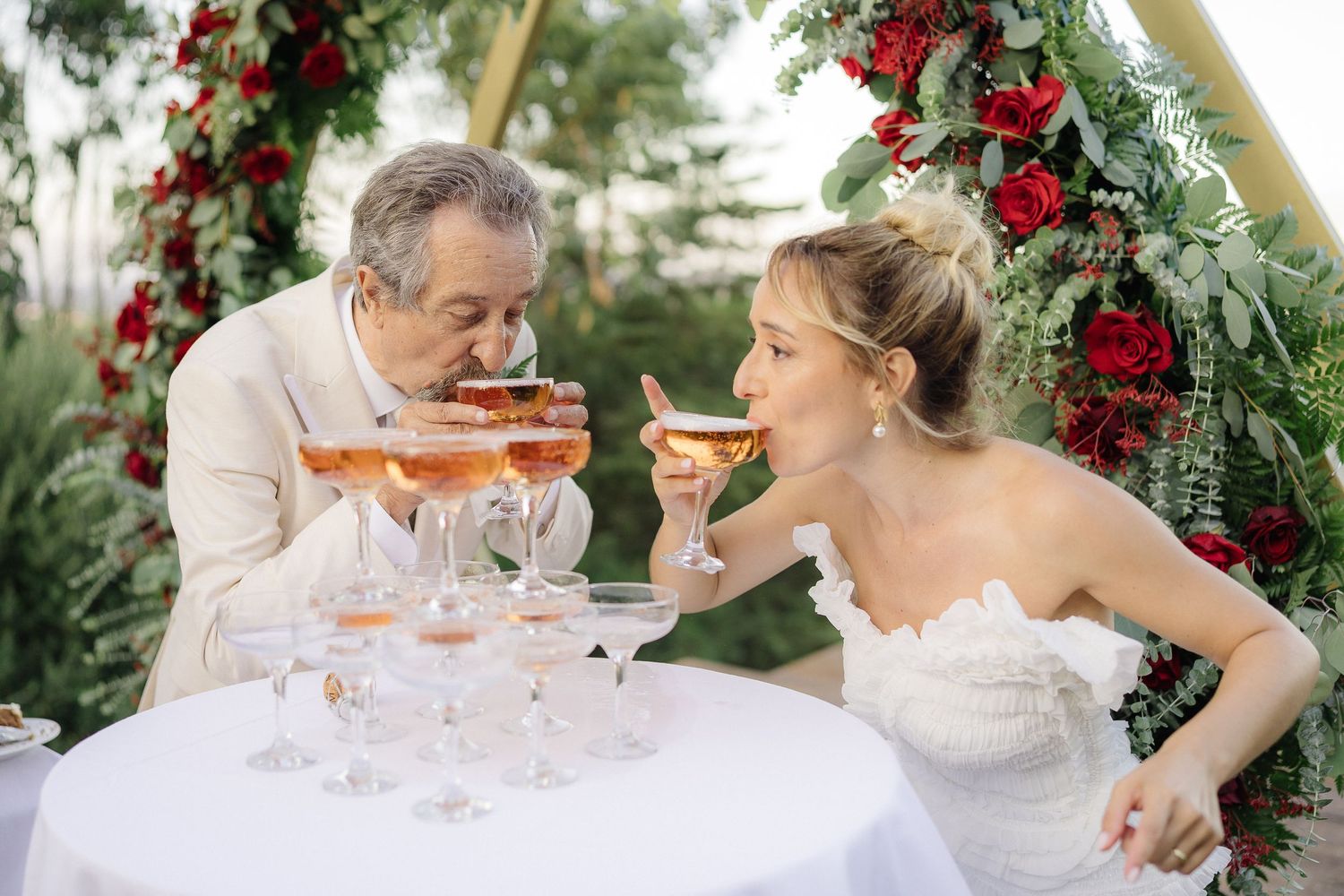 Couple sharing champagne under a floral arch decorated with red flowers.