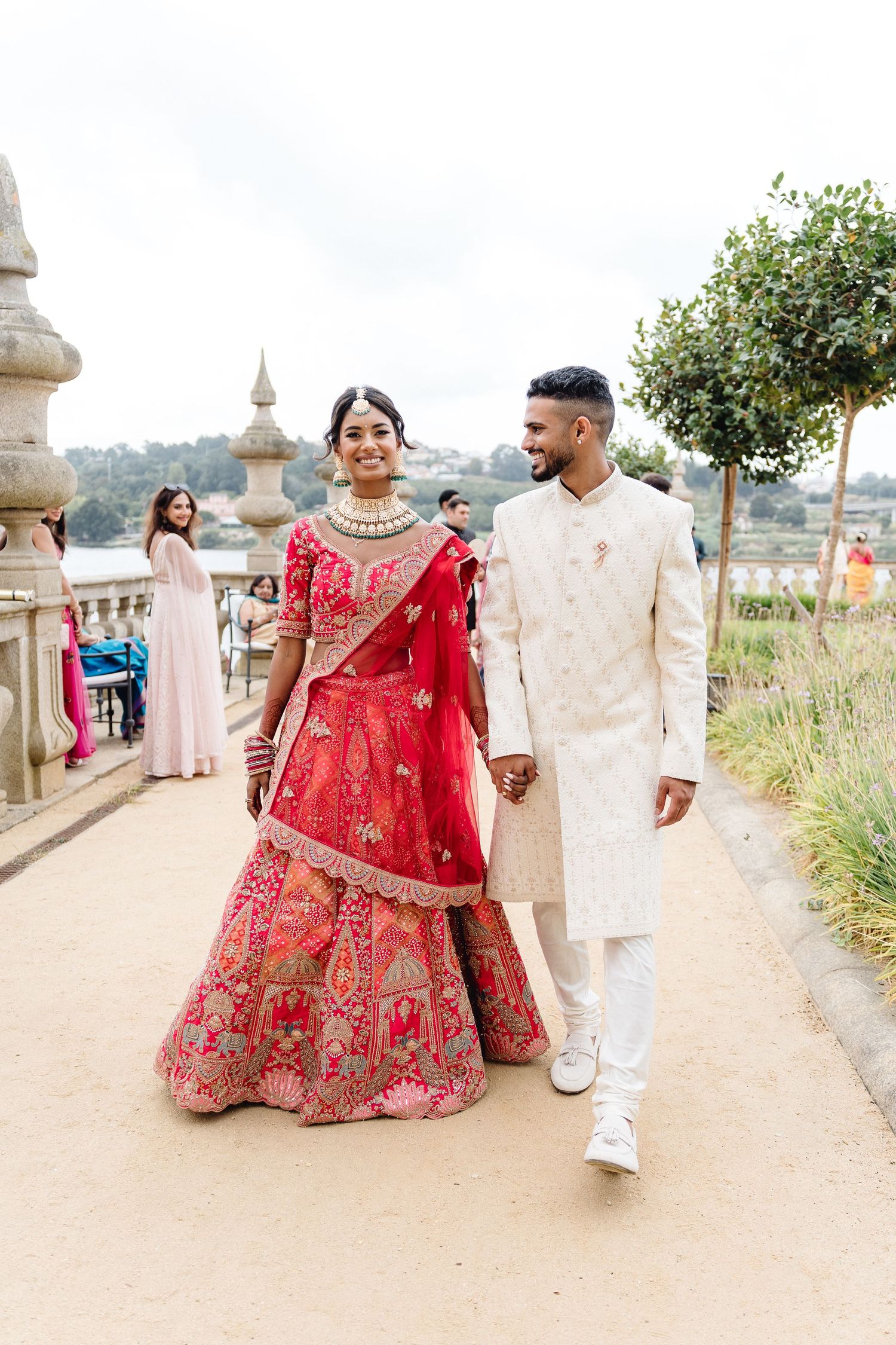 Couple in red lehenga and cream sherwani walking along outdoor pathway during wedding.