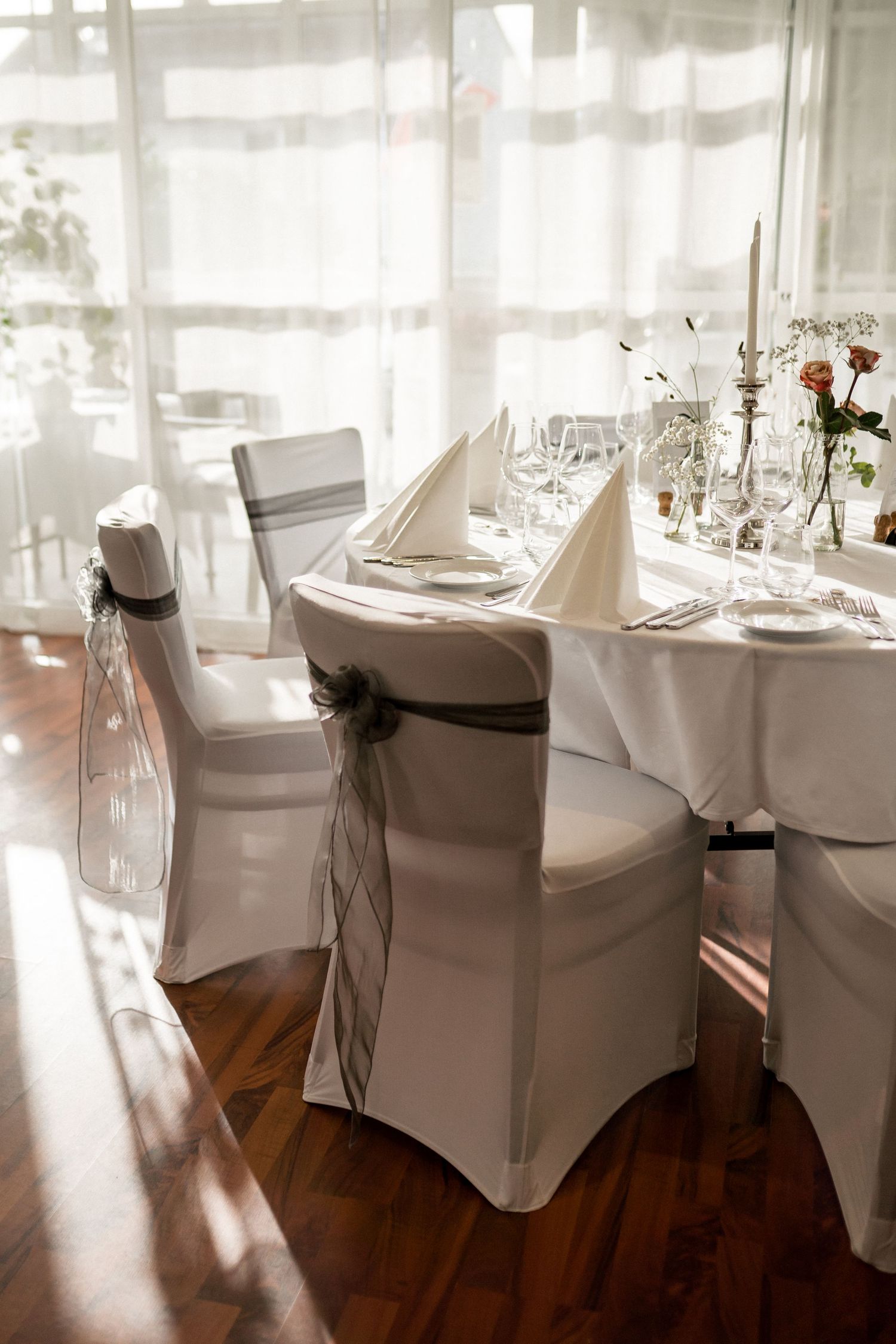 Elegant wedding reception tables set with white linens and chair covers in a bright sunlit room.
