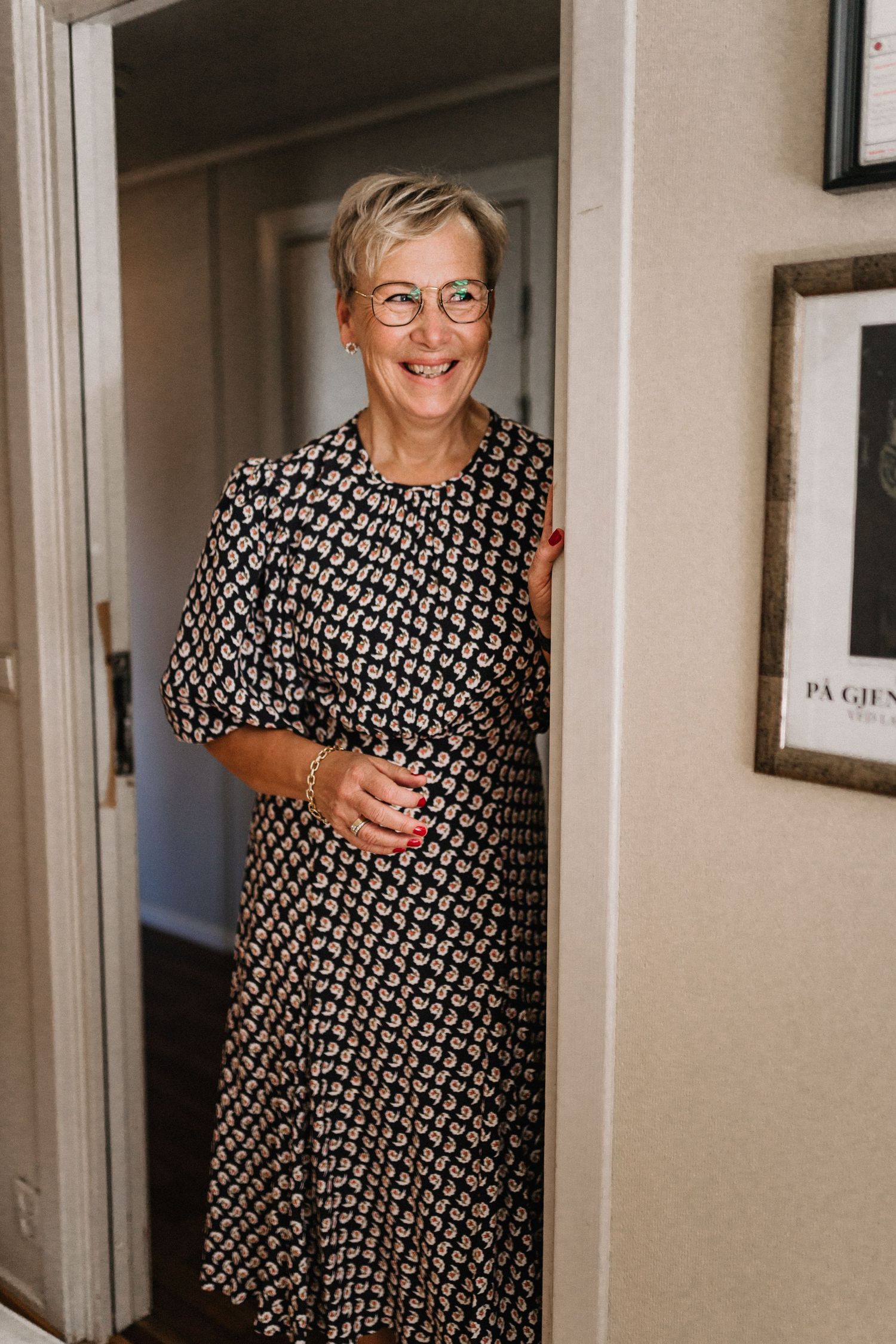 A figure in a black and white patterned dress stands in a hallway near framed artwork.