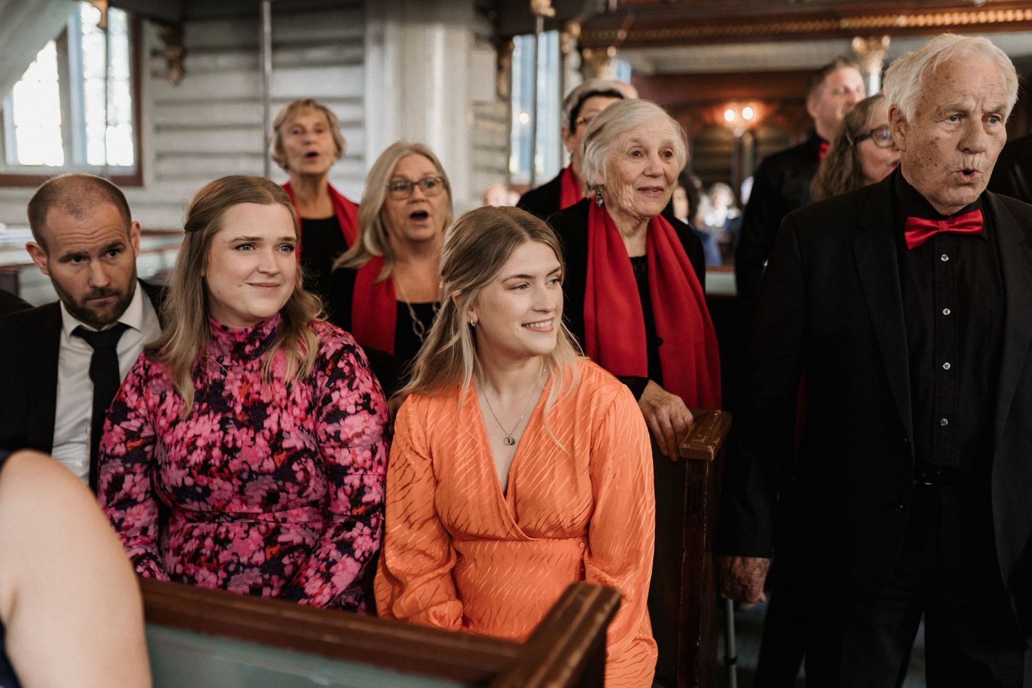Wedding guests in formal attire with pops of pink and orange clothing seated together.