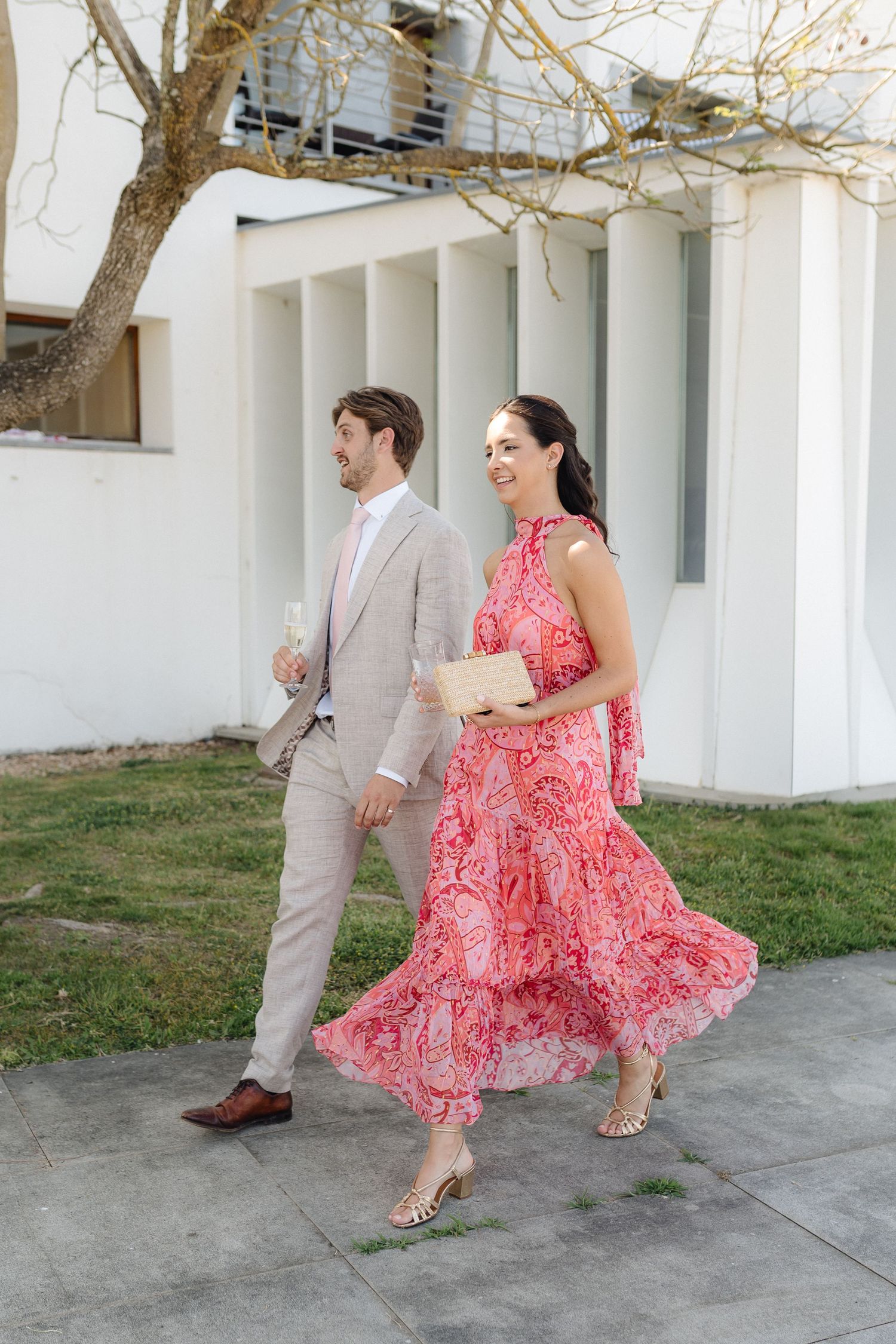 A couple walking together, with the woman wearing a flowing pink patterned dress and man in light beige suit.