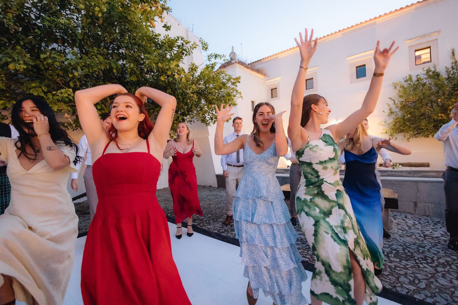 A group of wedding guests dancing joyfully together in colorful dresses at an outdoor celebration.