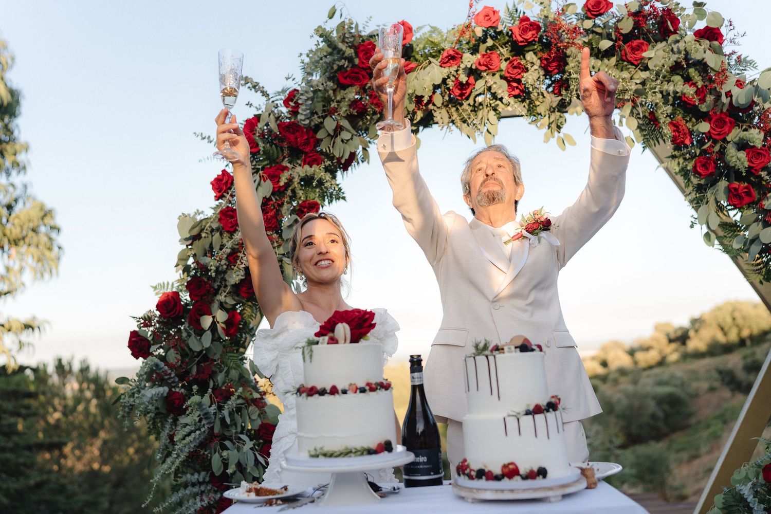 Wedding celebration under floral arch with red roses at golden hour.