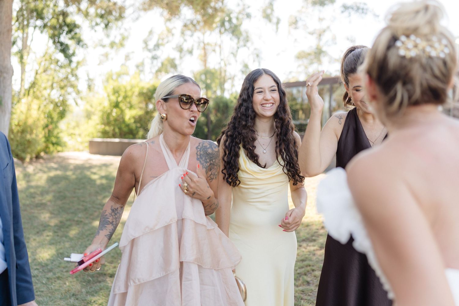 Guests in elegant dresses share laughter during an outdoor wedding celebration.