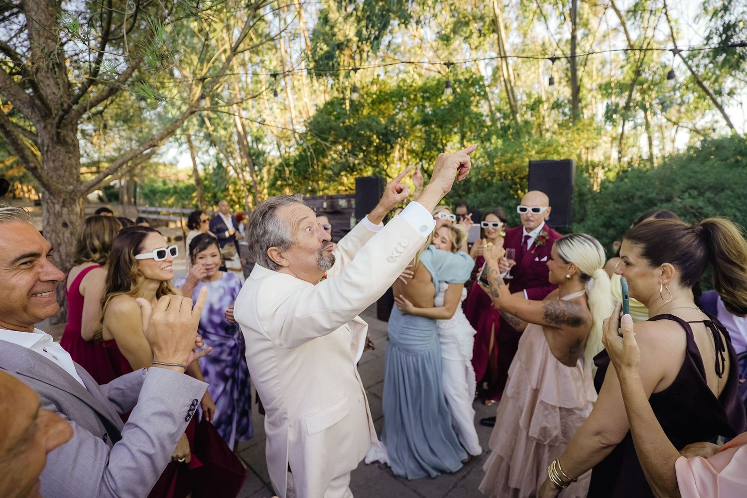 Large wedding party dances and celebrates outdoors under trees.