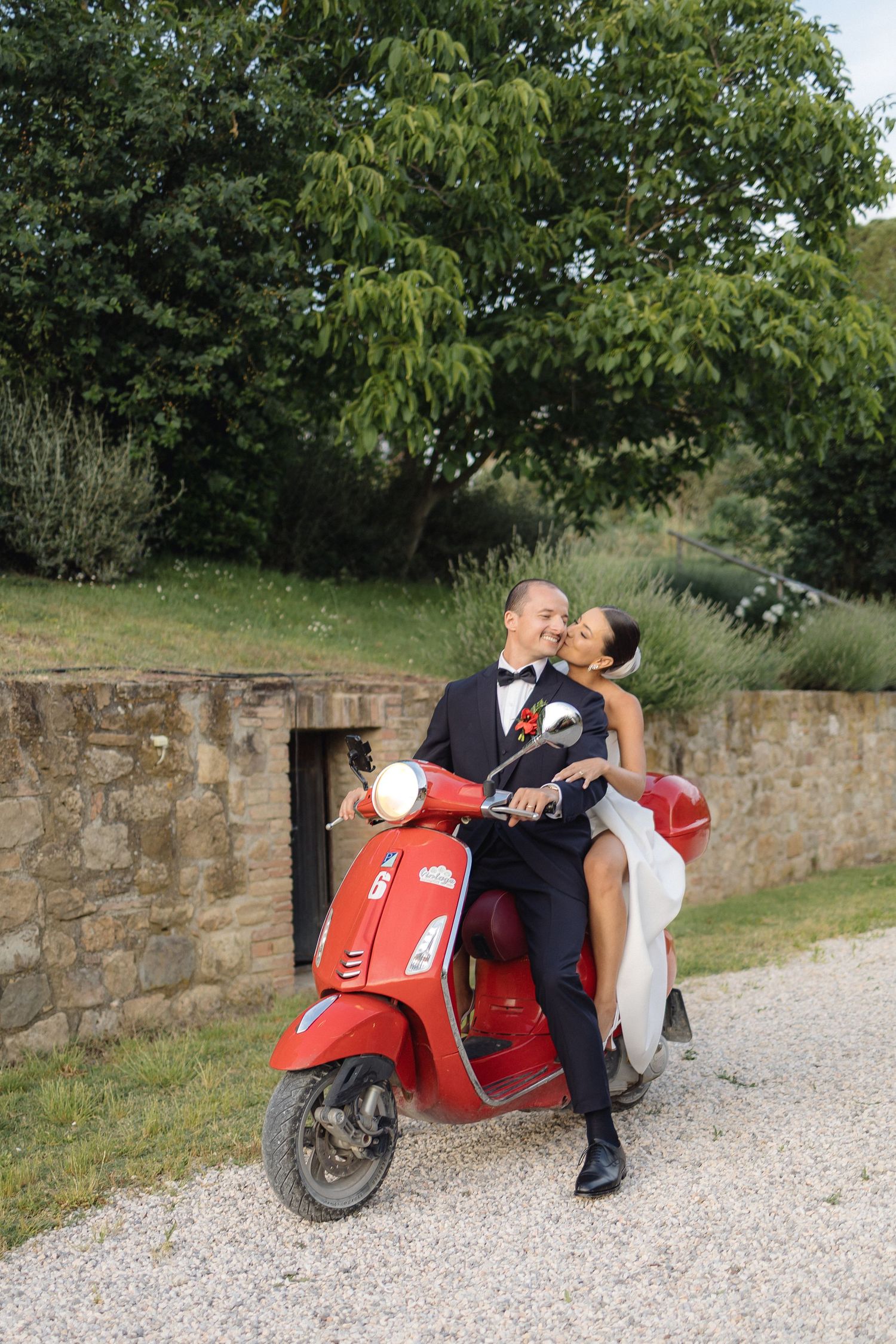 Bride and groom sharing a romantic kiss on a red Vespa scooter in a scenic Tuscan countryside setting.