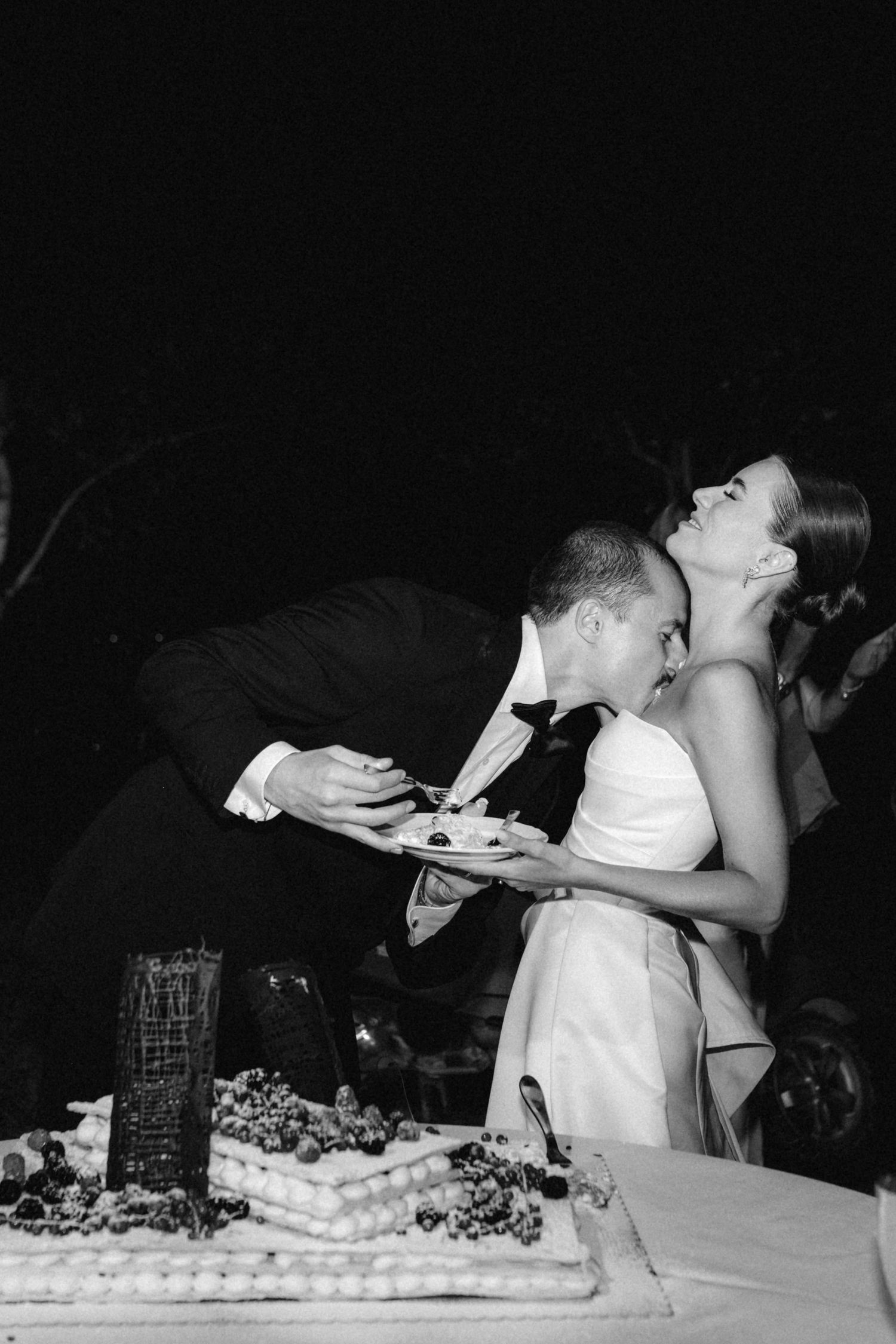 Bride and groom sharing wedding cake in playful black and white nighttime reception photograph.