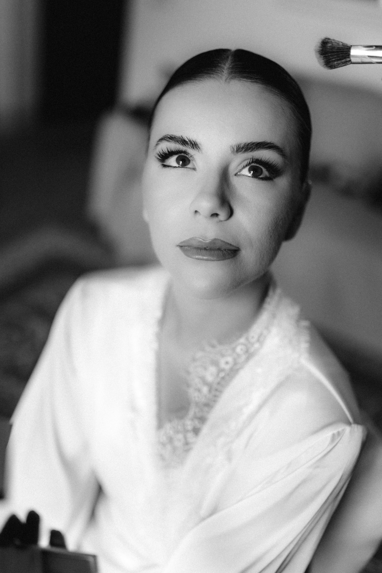 Black and white portrait of a bride getting makeup applied with a brush during wedding preparations.