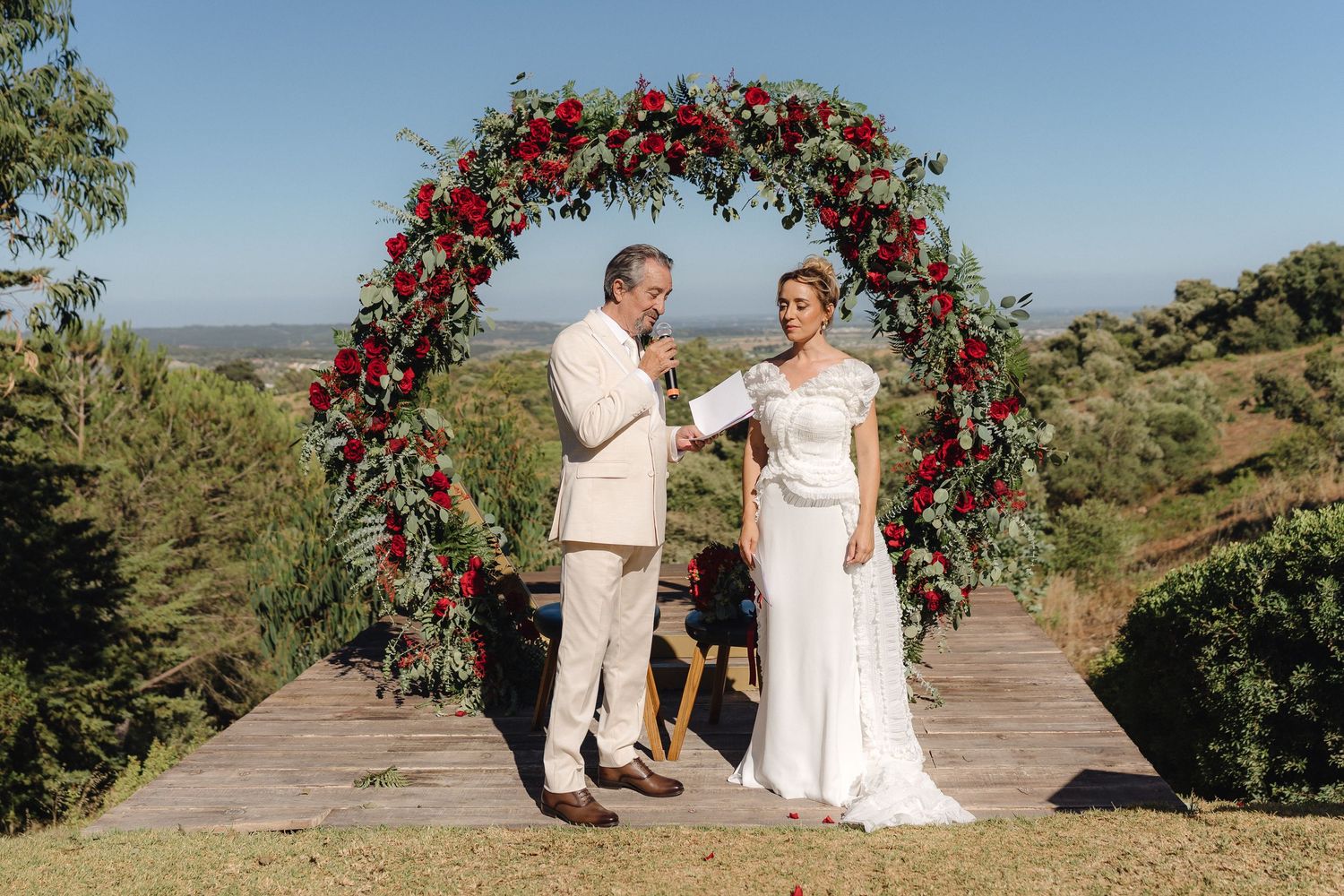 A romantic wedding ceremony takes place under an arch adorned with red roses on a scenic overlook.