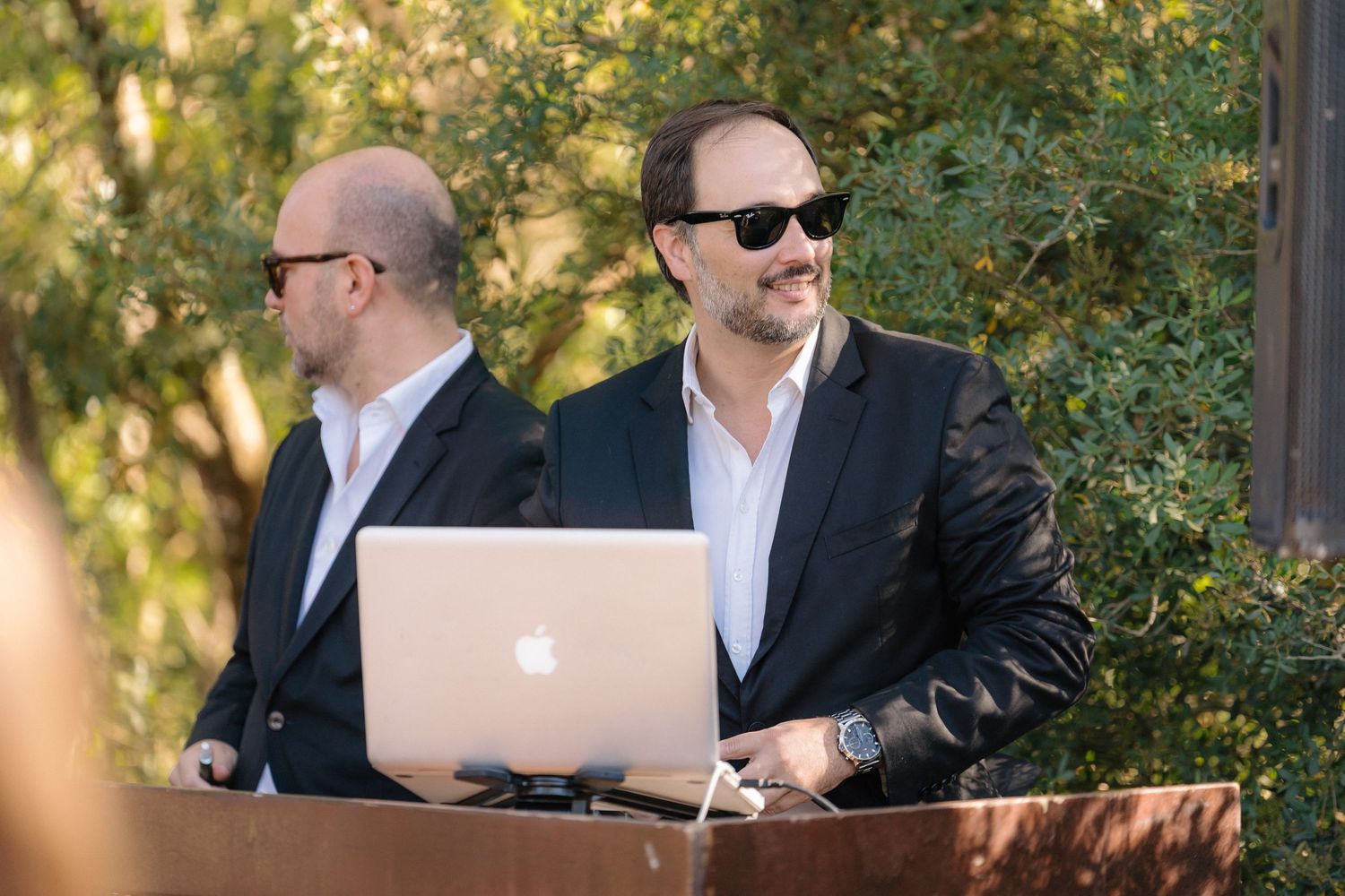 DJs in suits operate equipment with a laptop at an outdoor event.