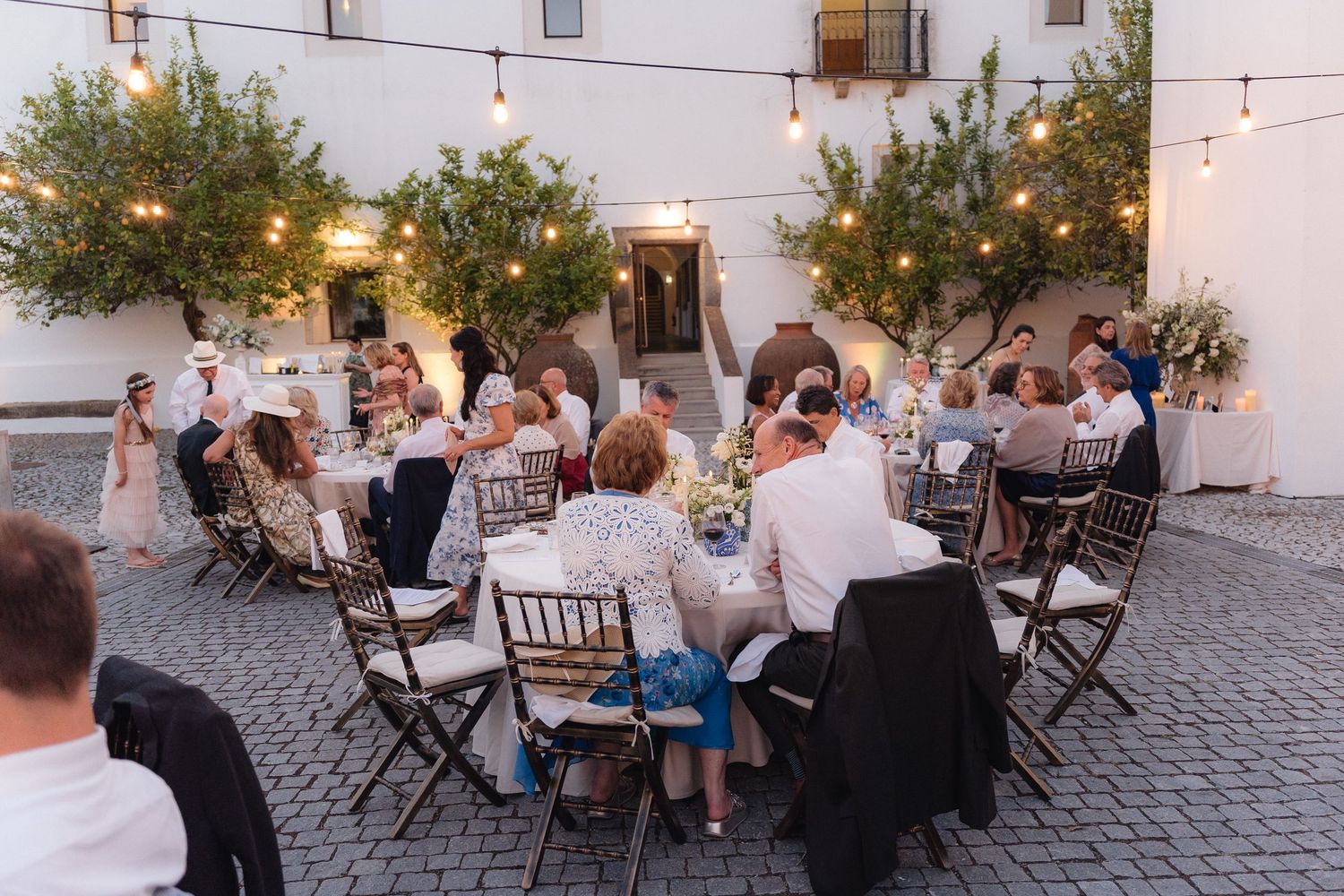 Evening outdoor reception with guests seated at tables under string lights and olive trees.