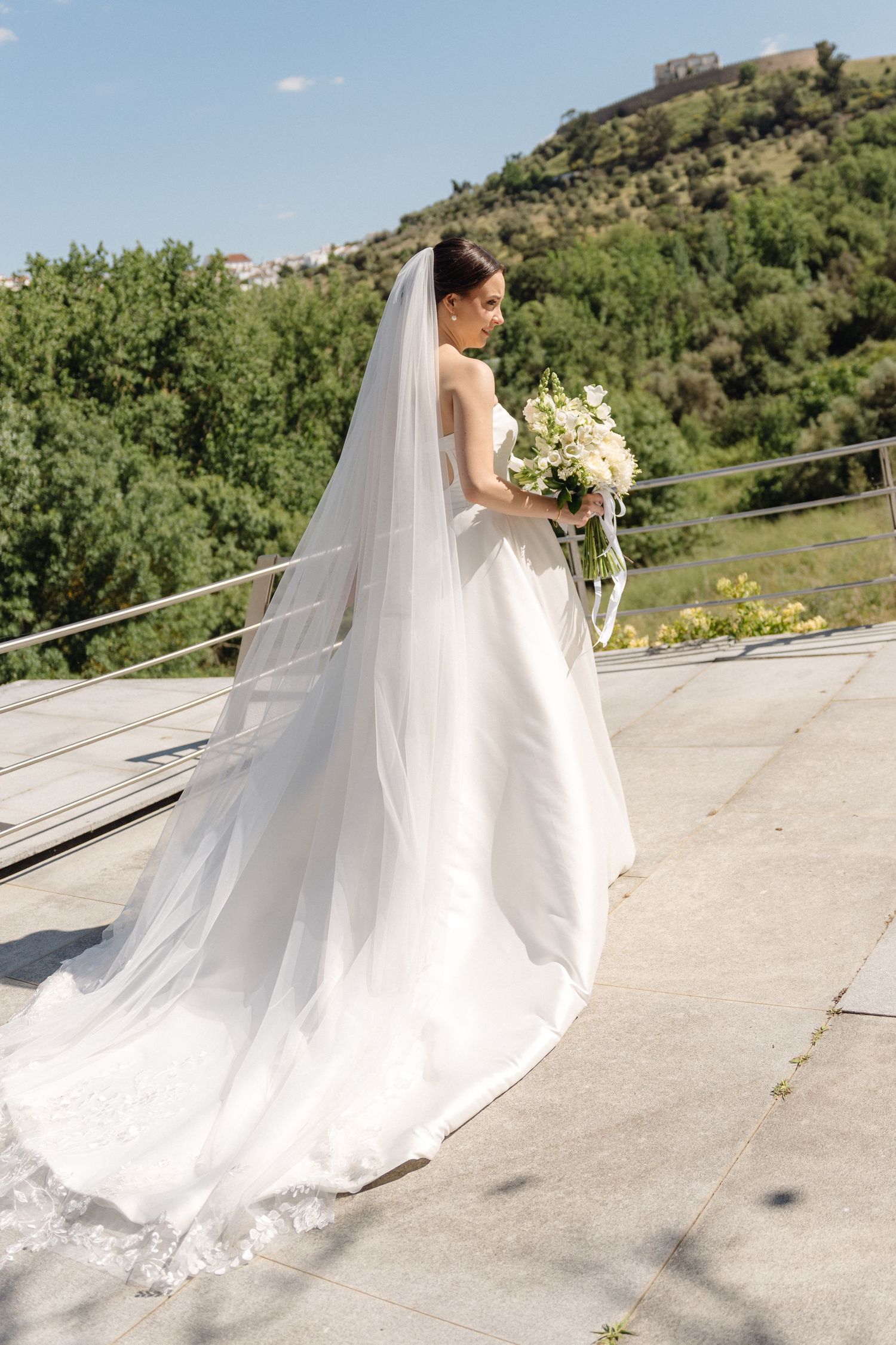 Flowing white wedding dress with long train photographed on outdoor terrace.
