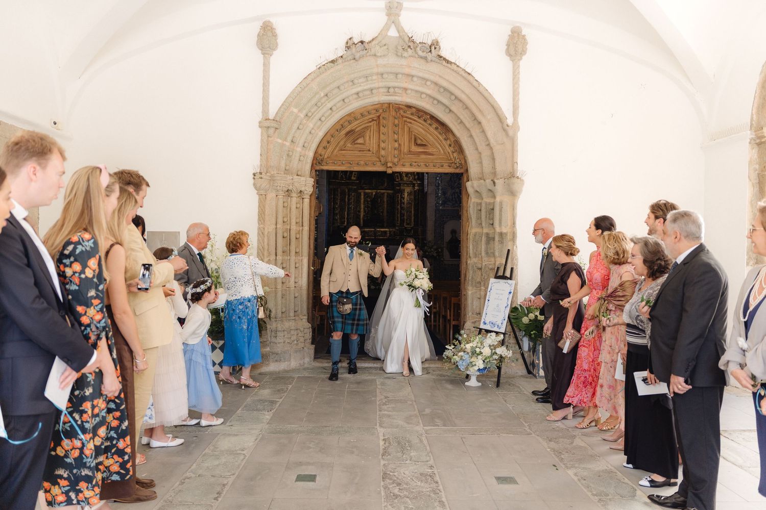 Wedding party and guests mingling in historic church corridor with arched ceiling.