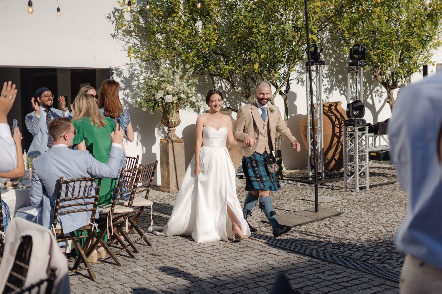 Wedding ceremony guests seated in rows on cobblestone courtyard with white church walls.