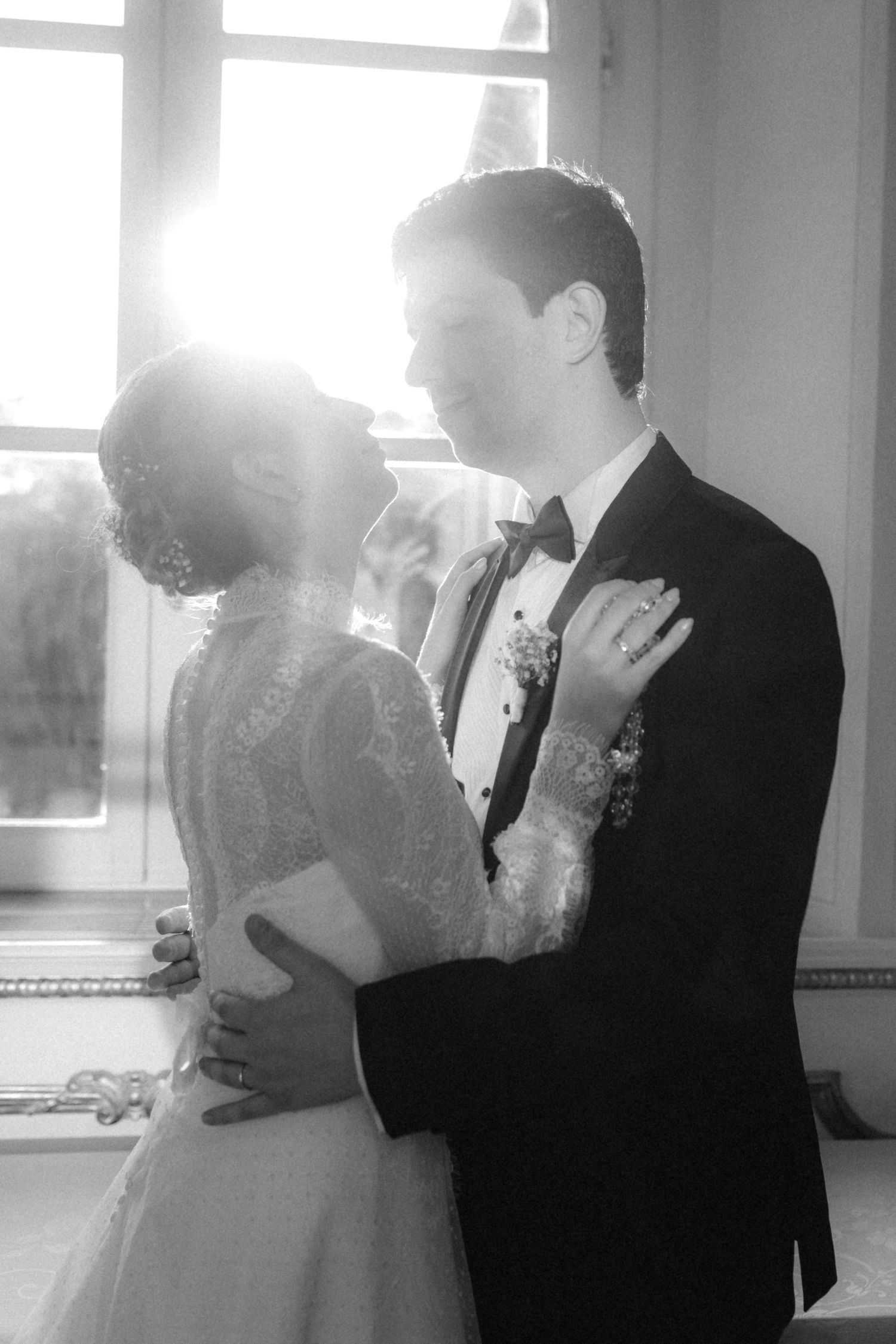 Black and white photo of bride and groom embracing near sunlit window in elegant wedding attire.