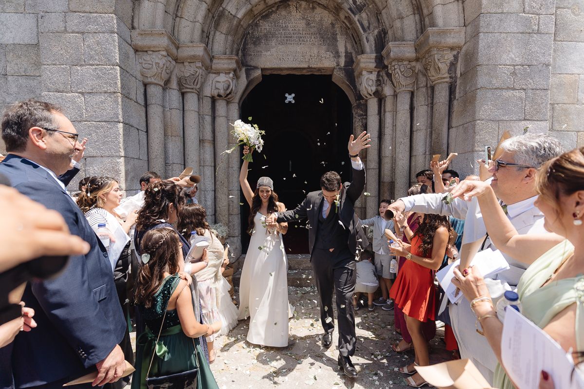 Wedding party exits historic stone church as guests celebrate with flower petals.