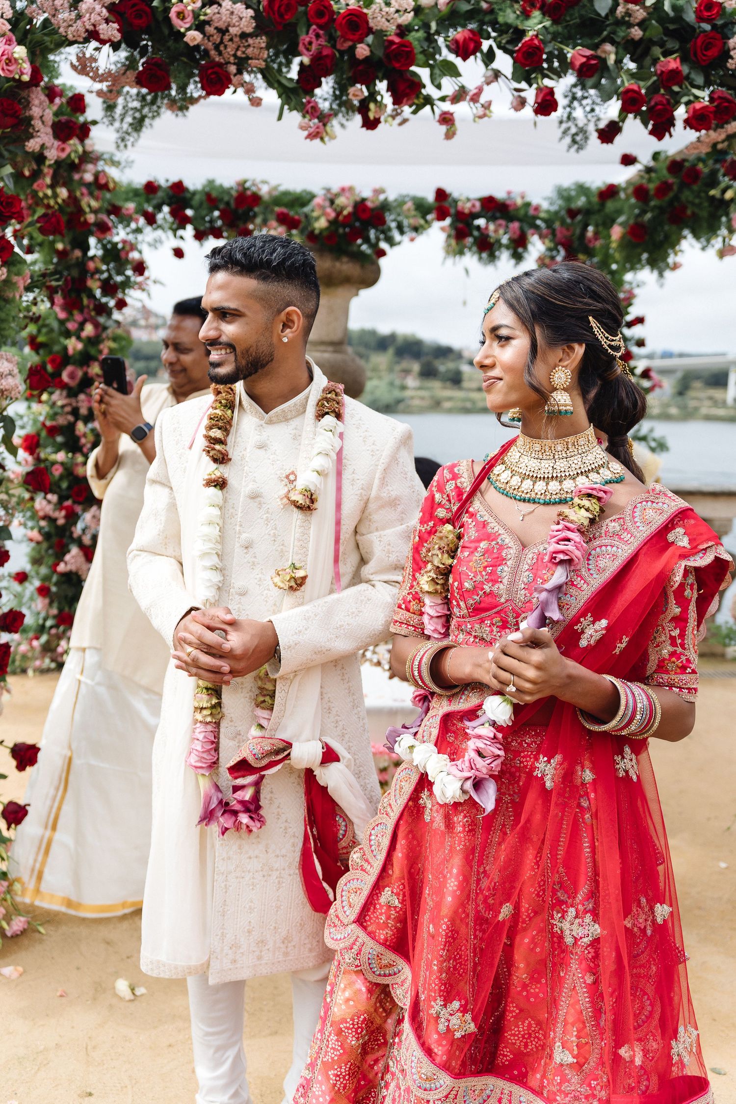 Couple stands under floral arch with red roses wearing traditional Indian wedding attire.