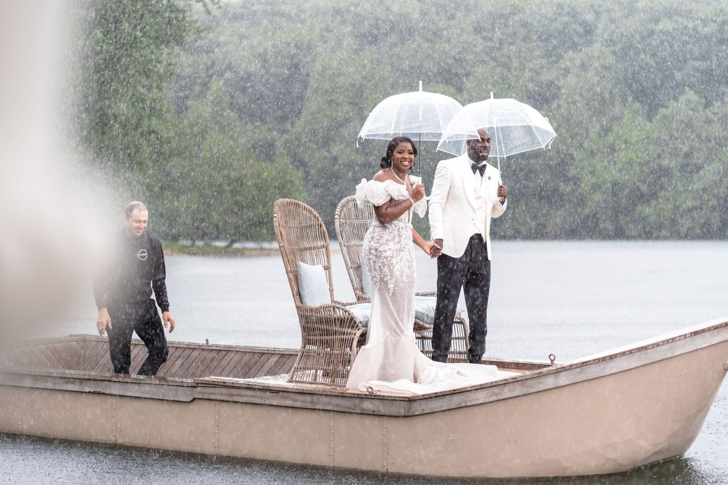 Wedding party members with umbrellas gather near a lake on an overcast day.