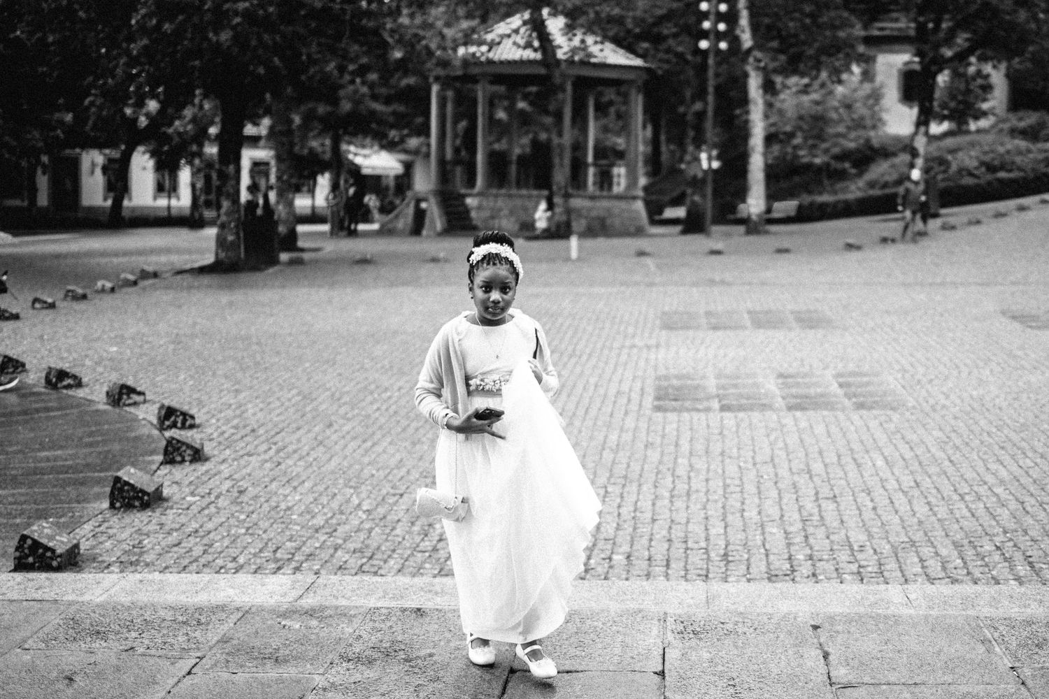 A flower girl in white walks down a cobblestone path during a wedding ceremony.
