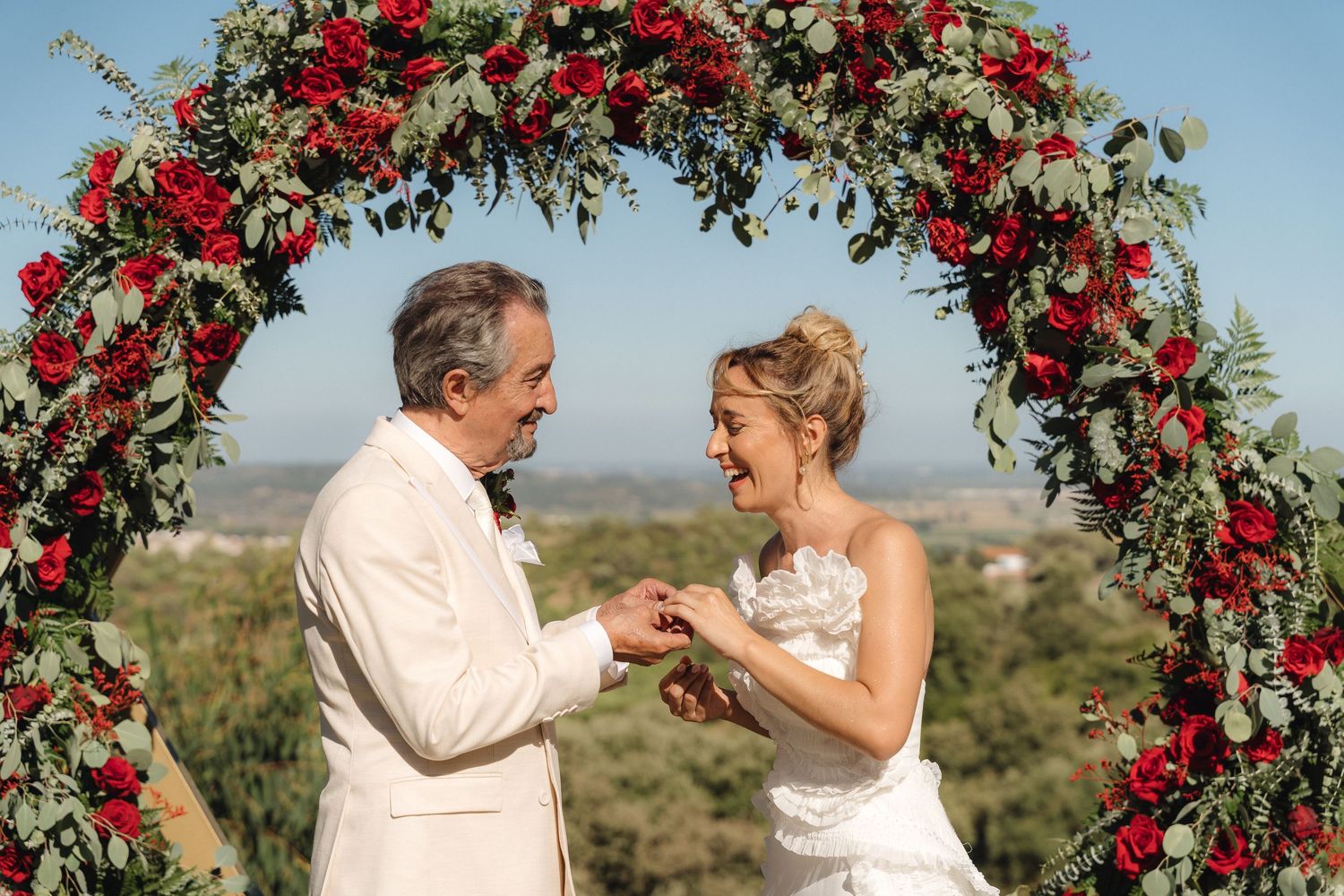 Intimate wedding moment captured under a lush archway of red roses against blue skies.
